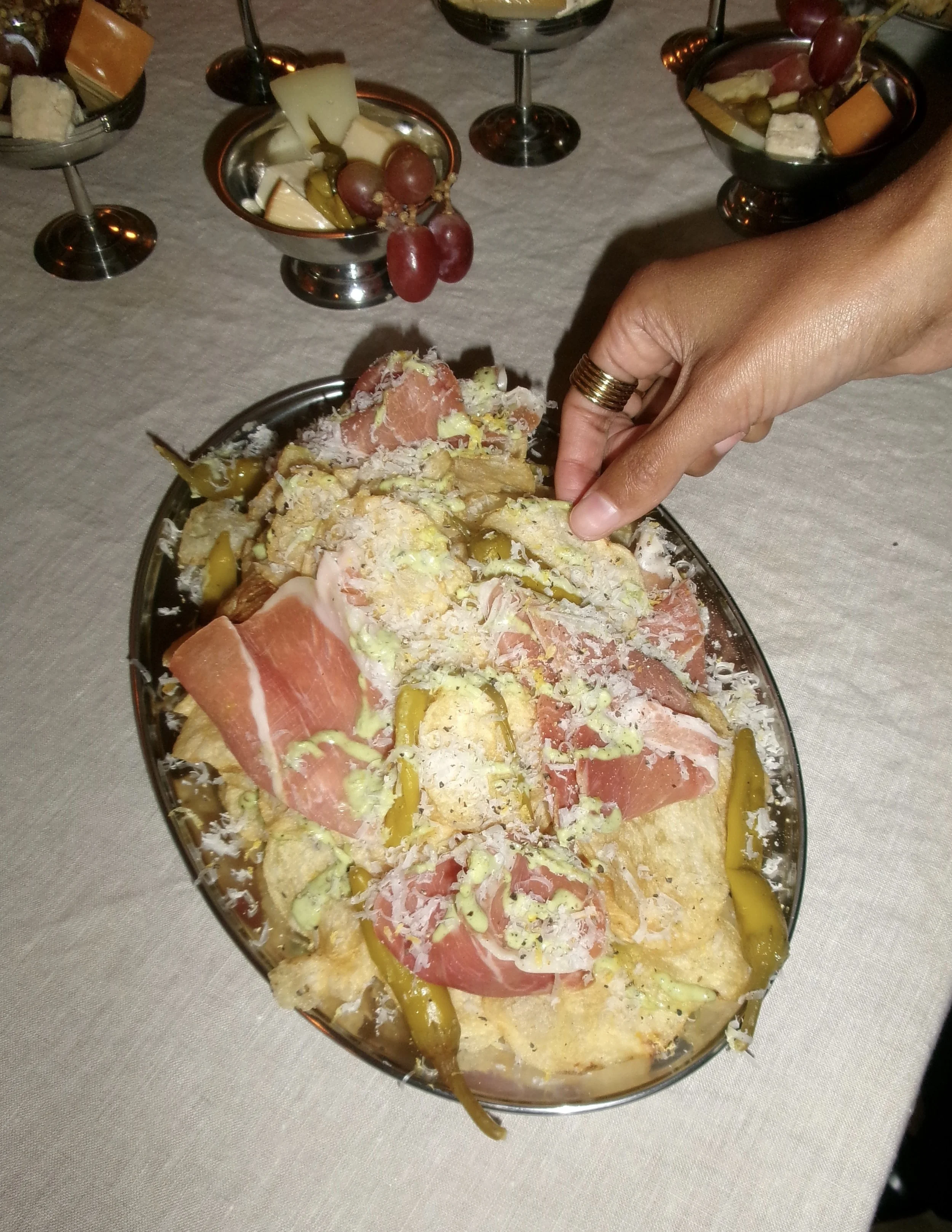 Person with a ring reaching for a dish of layered, yellow potato chips topped with prosciutto, pickles, cheese, and sauce, with small glass bowls of cheese, grapes, and pickles on a table in the background.