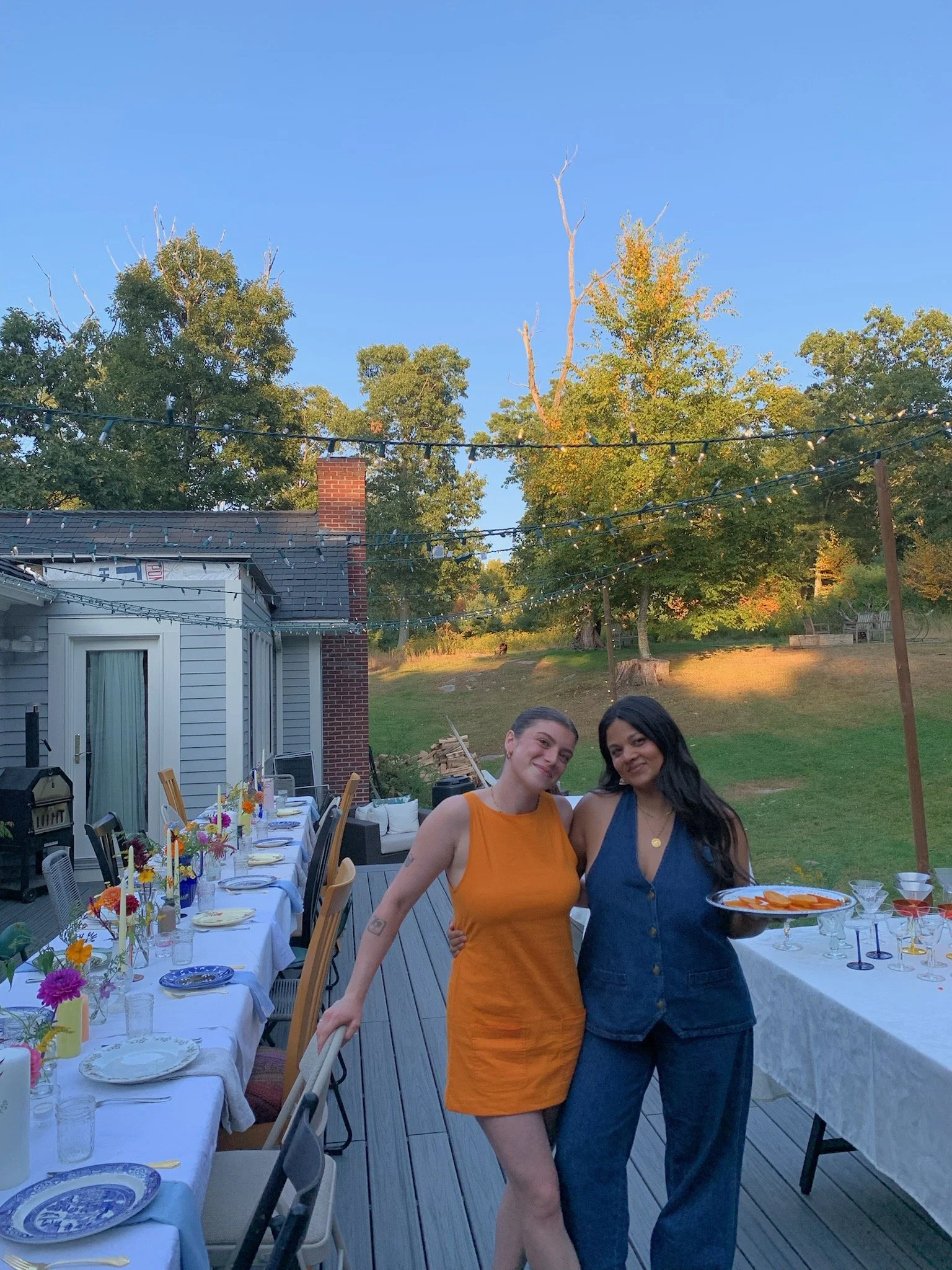Two women standing together, smiling at a backyard dinner party, with tables set for a meal, string lights, and trees in the background during sunset.