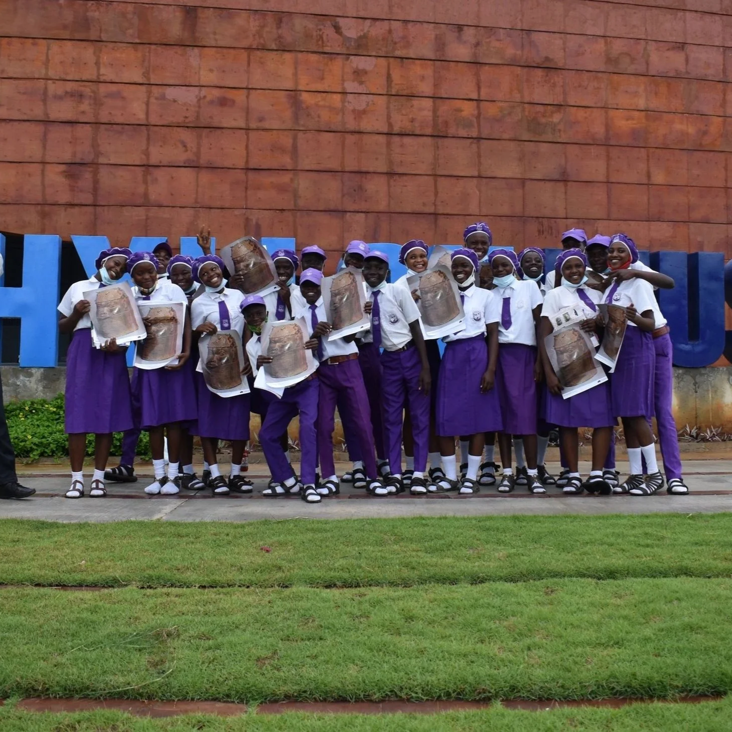 Group of school children wearing white shirts and purple skirts or shorts, holding certificates, standing outdoors in front of a building with a blue sign.