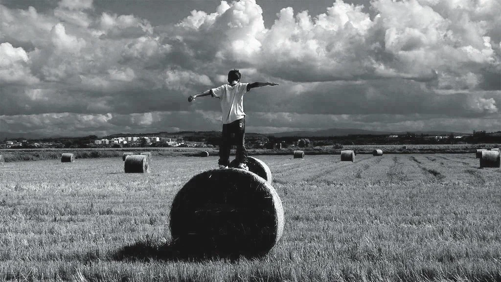 Person standing on top of a large hay bale in a field with multiple hay bales scattered around, under a cloudy sky.