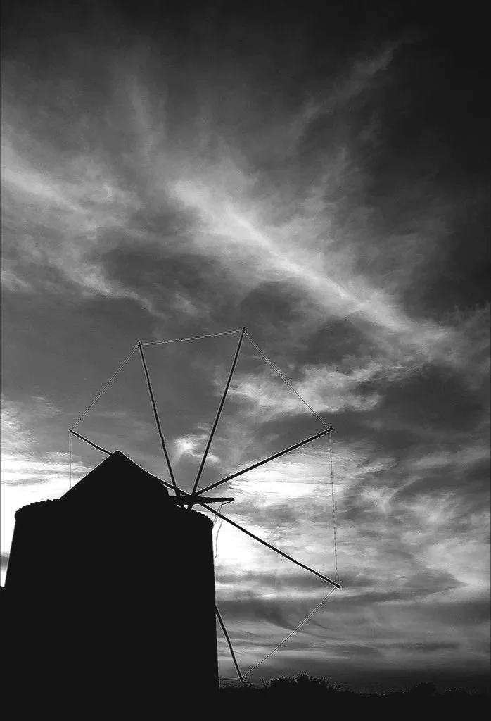 Silhouette of a windmill or antenna on top of a building against a cloudy sky during sunset or sunrise.