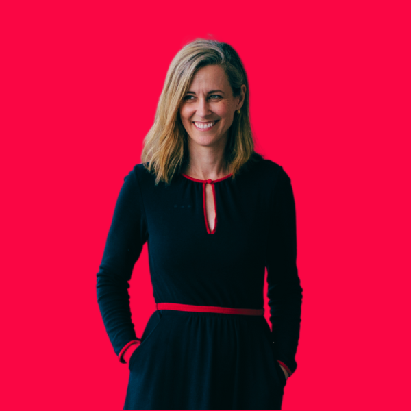 A woman with shoulder-length blonde hair, wearing a black dress with red accents, smiling and standing against a bright pink background.