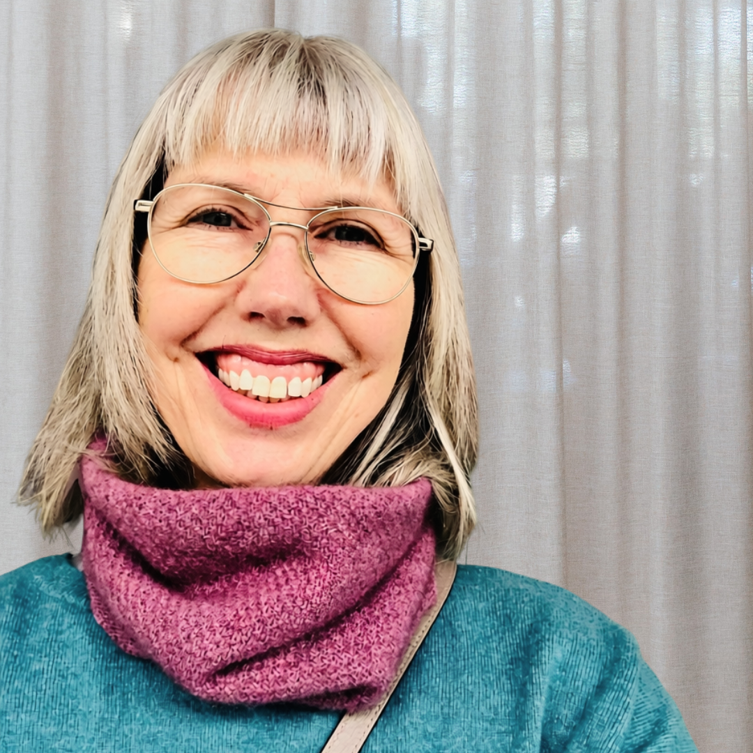 A smiling woman with glasses, blonde hair with dark streaks, wearing a teal top and a pink scarf standing in front of a light-colored curtain.