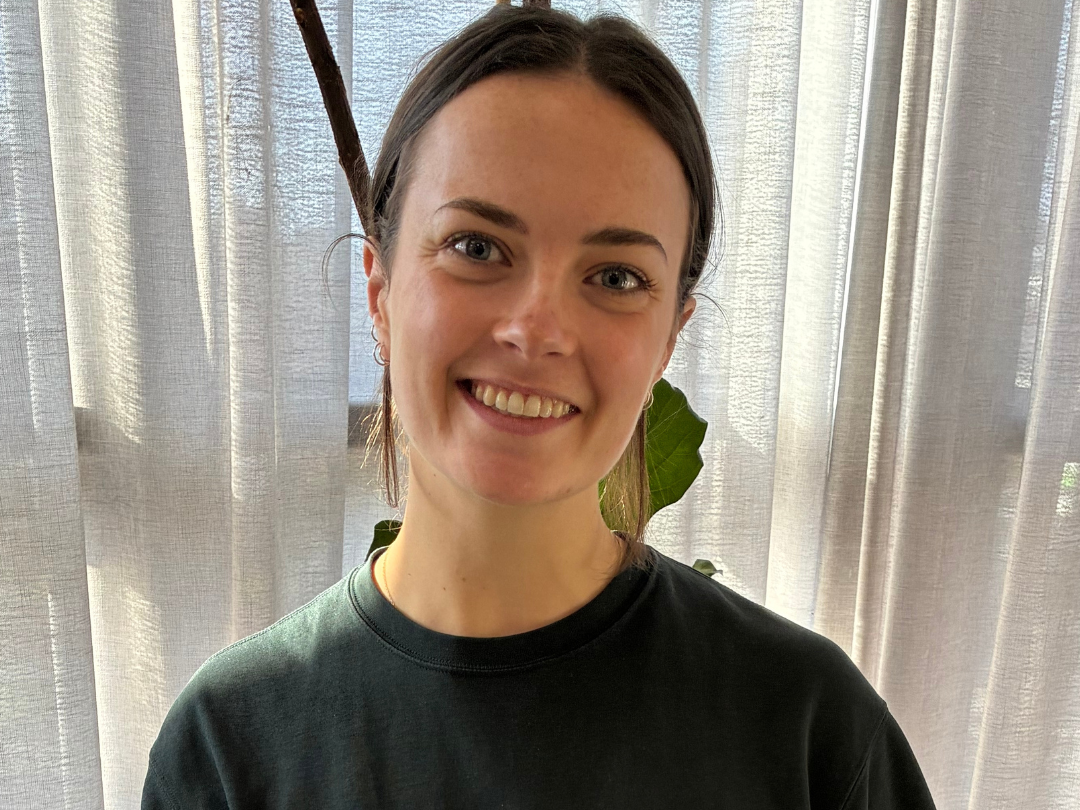 Young woman with short dark hair, wearing a white shirt, smiling, standing near a large green plant with broad leaves and sheer curtains in the background.