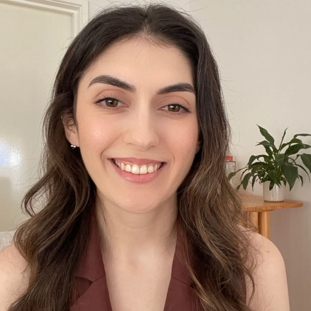 A woman smiling indoors with long wavy hair, wearing a brown top. There's a green plant on a wooden shelf in the background.