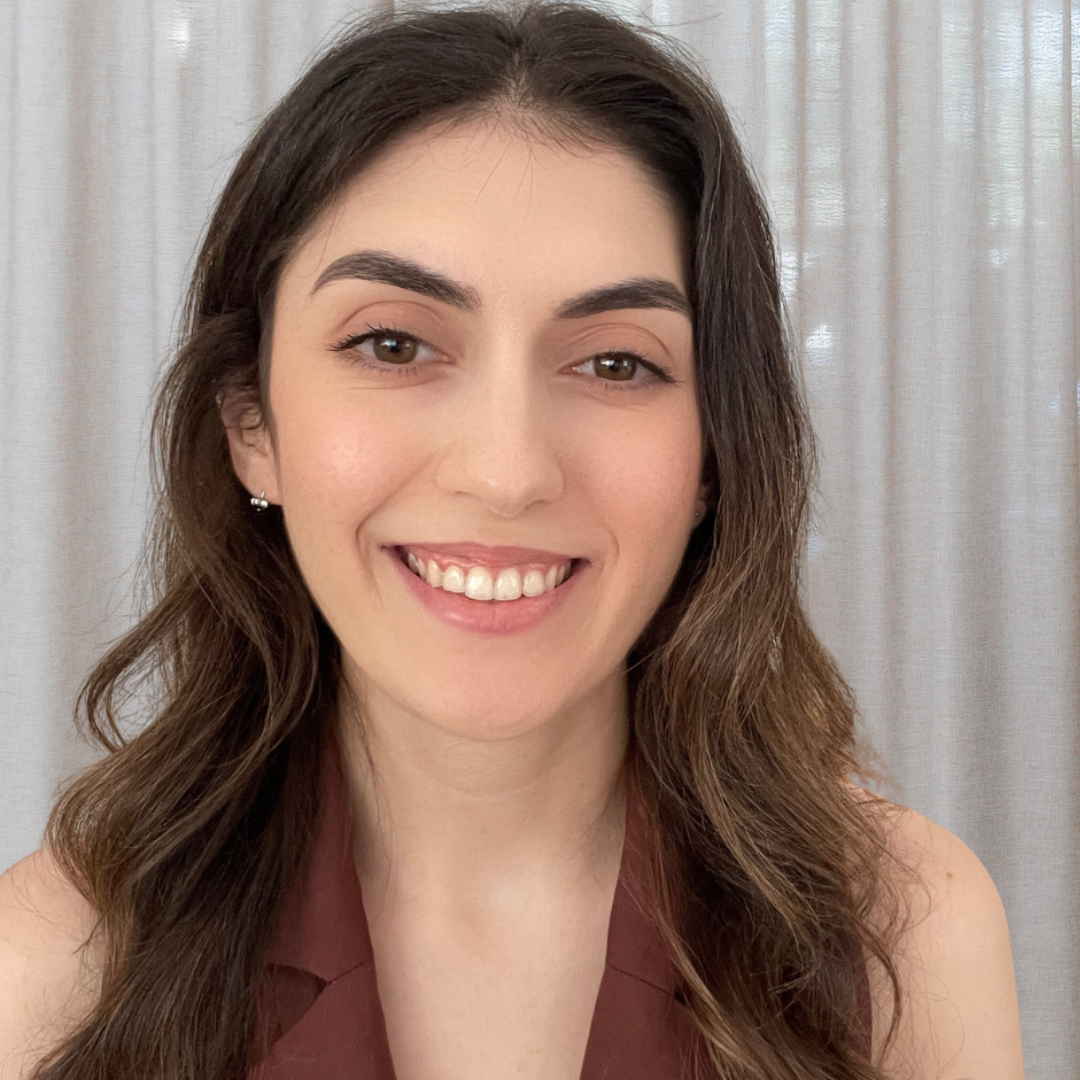 A woman smiling indoors with long wavy hair, wearing a brown top. There's a green plant on a wooden shelf in the background.