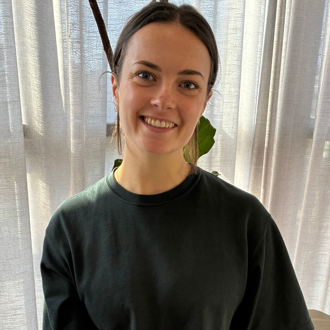 Person wearing a white T-shirt smiling, standing indoors with a plant and sheer curtains in the background.
