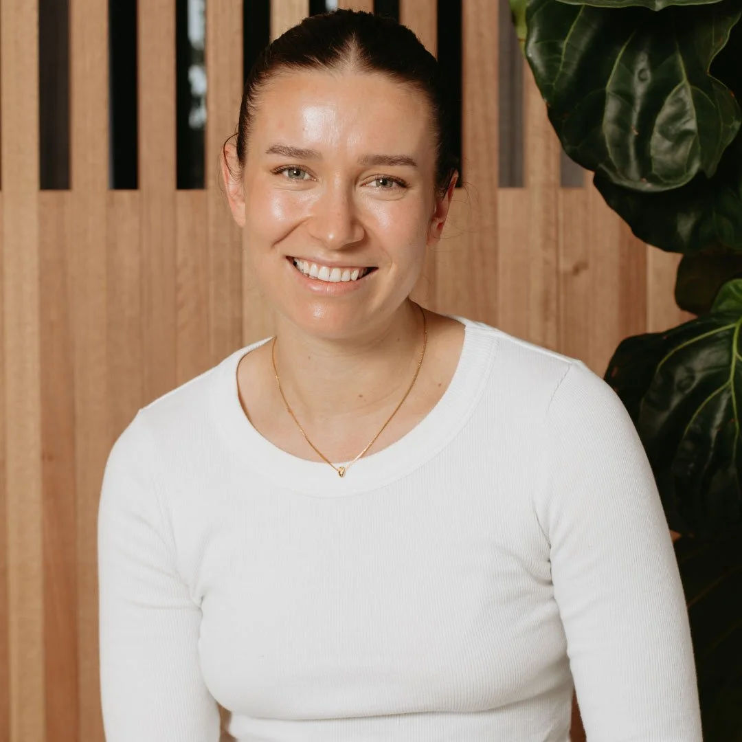 Person wearing a white shirt and necklace smiling in front of wooden paneling and plant.