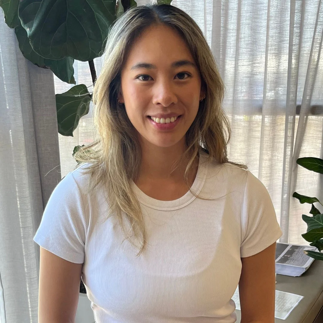 A woman with long blonde hair wearing a white shirt, smiling indoors with plants in the background.