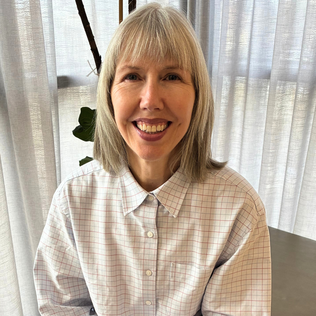A smiling woman with blonde hair wearing a button up white checkerd shirt, sitting in front of gray curtains.