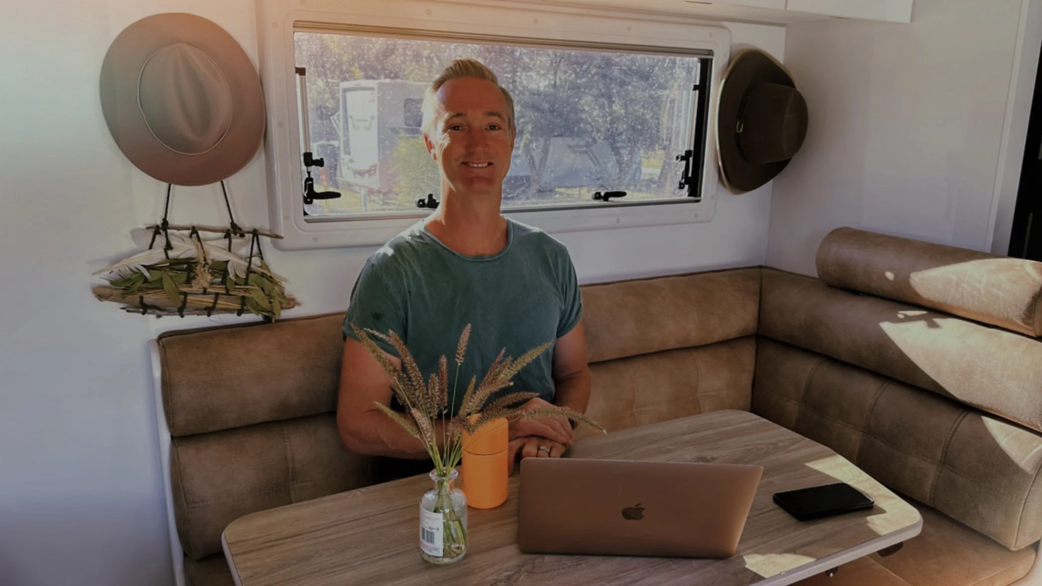 Person sitting at a table in an RV, with a laptop, vase of grass, and a mug on the table. Two hats are hanging on the nearby wall, and sunlight is coming through the window.