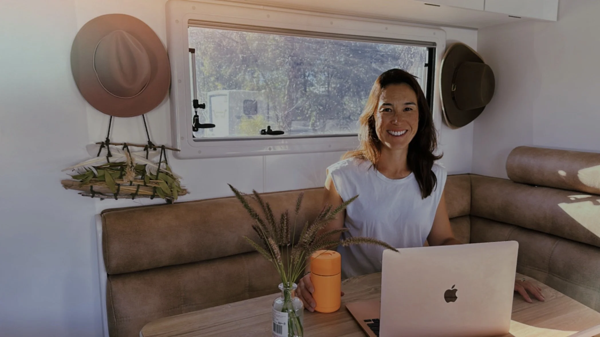 Woman sitting at a table inside a camper, smiling, with a laptop, mug, and vase with plants on the table, hats hanging on the wall.
