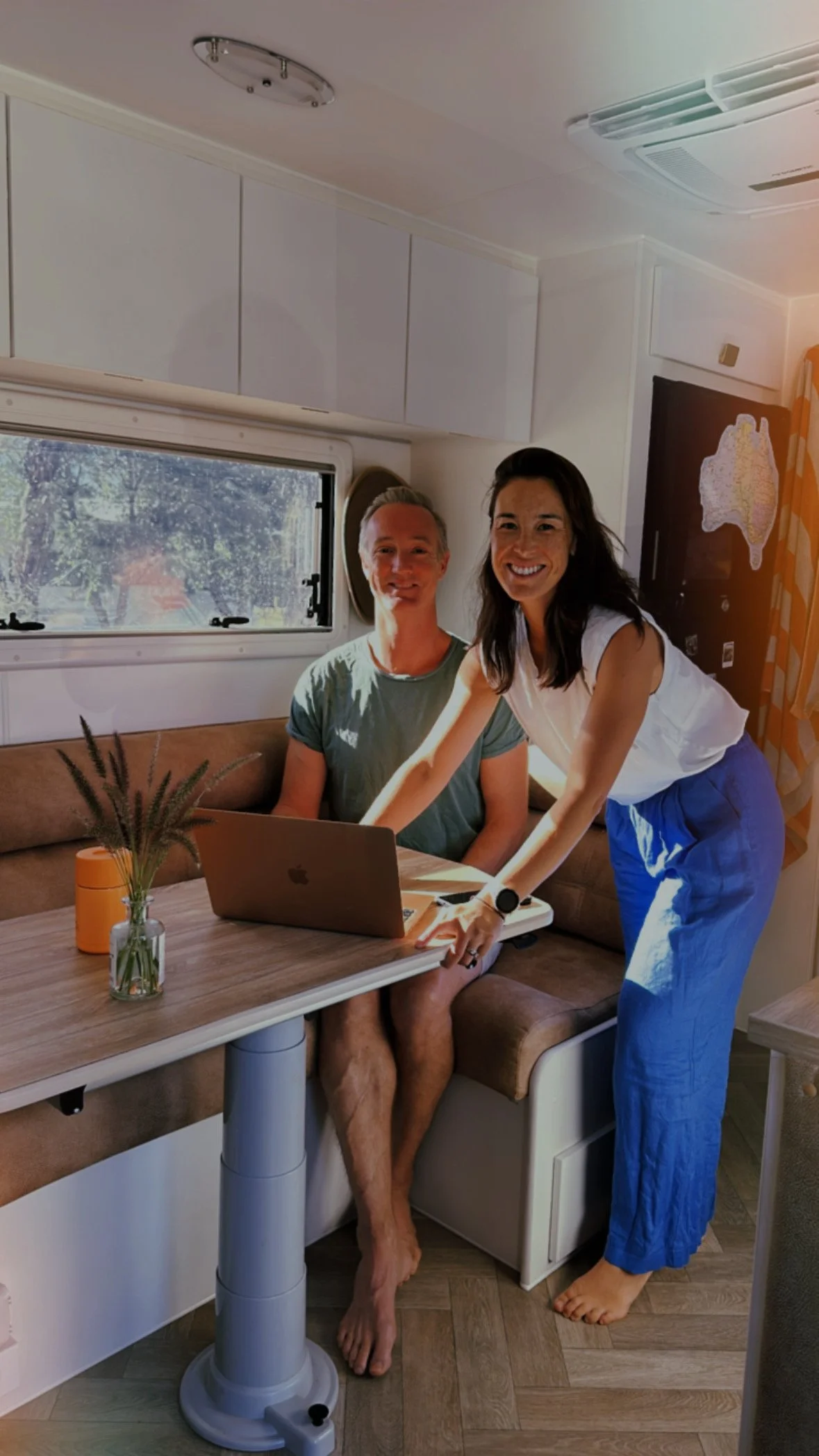 Two people inside a camper van, one sitting with a laptop and another standing, both smiling. The interior includes a table, window, and cabinets.