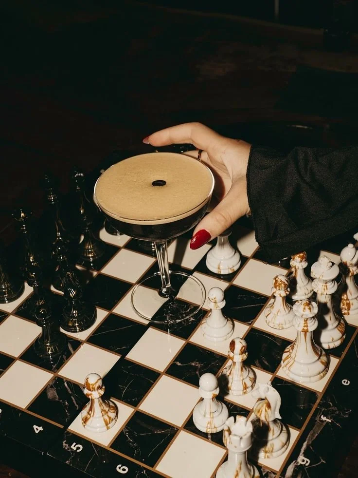 A hand with red painted nails holding a cocktail glass with a dark brown drink on a black and white chessboard table.