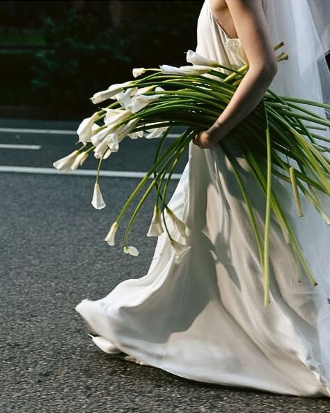 Bride in white dress holding large bouquet of calla lilies, walking on pavement.