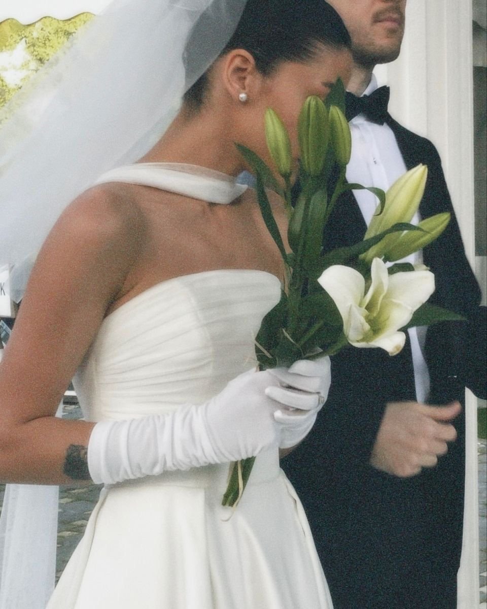 A bride holding white lilies, standing next to a groom in a tuxedo, with her face partially covered by the flowers and veiled, during a wedding ceremony.