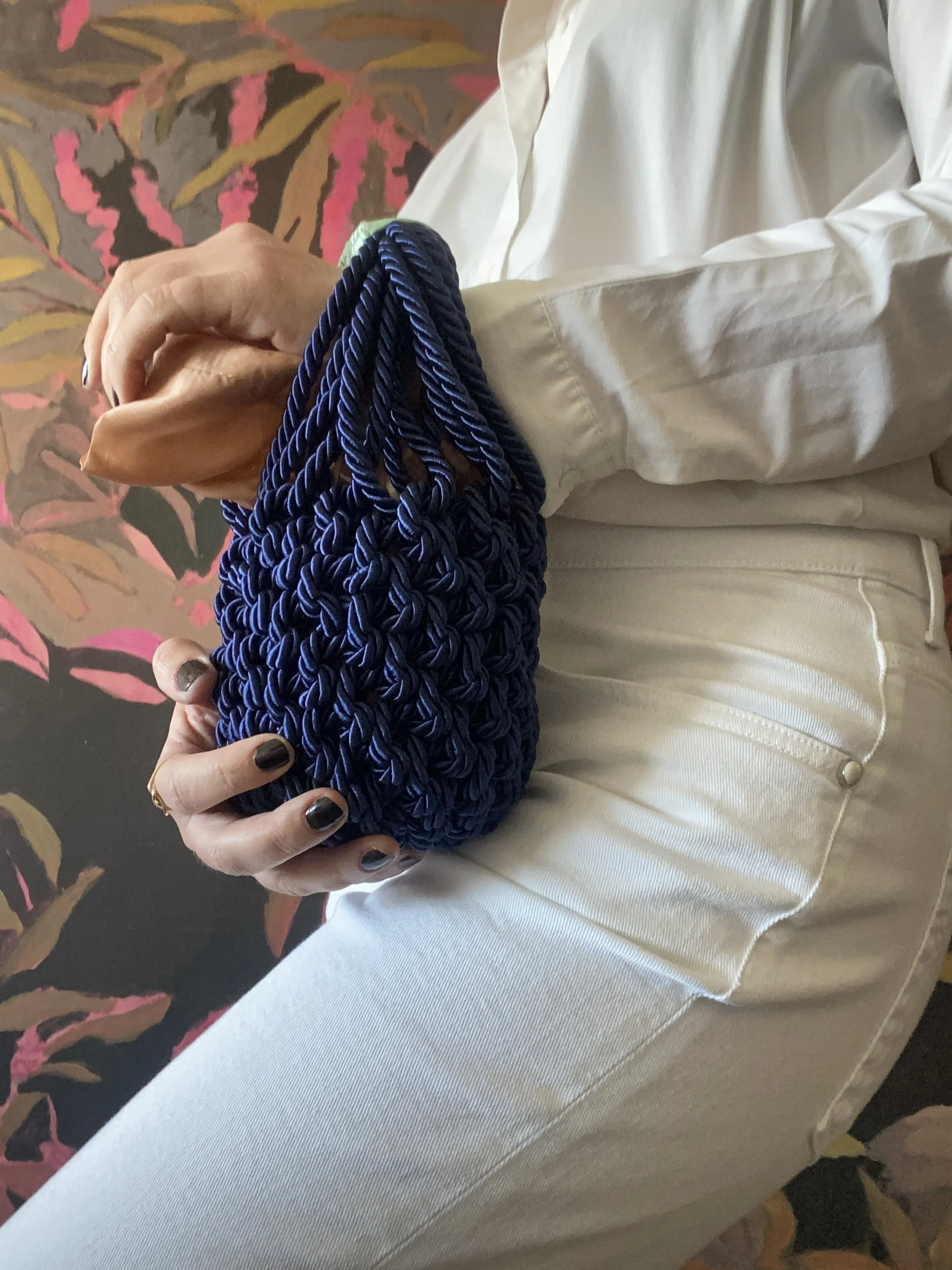Una persona tiene in mano una borsa blu a tessitura intrecciata, indossa pantaloni bianchi e una camicia bianca, sfondo con motivo di foglie rosa, gialle e marroni.