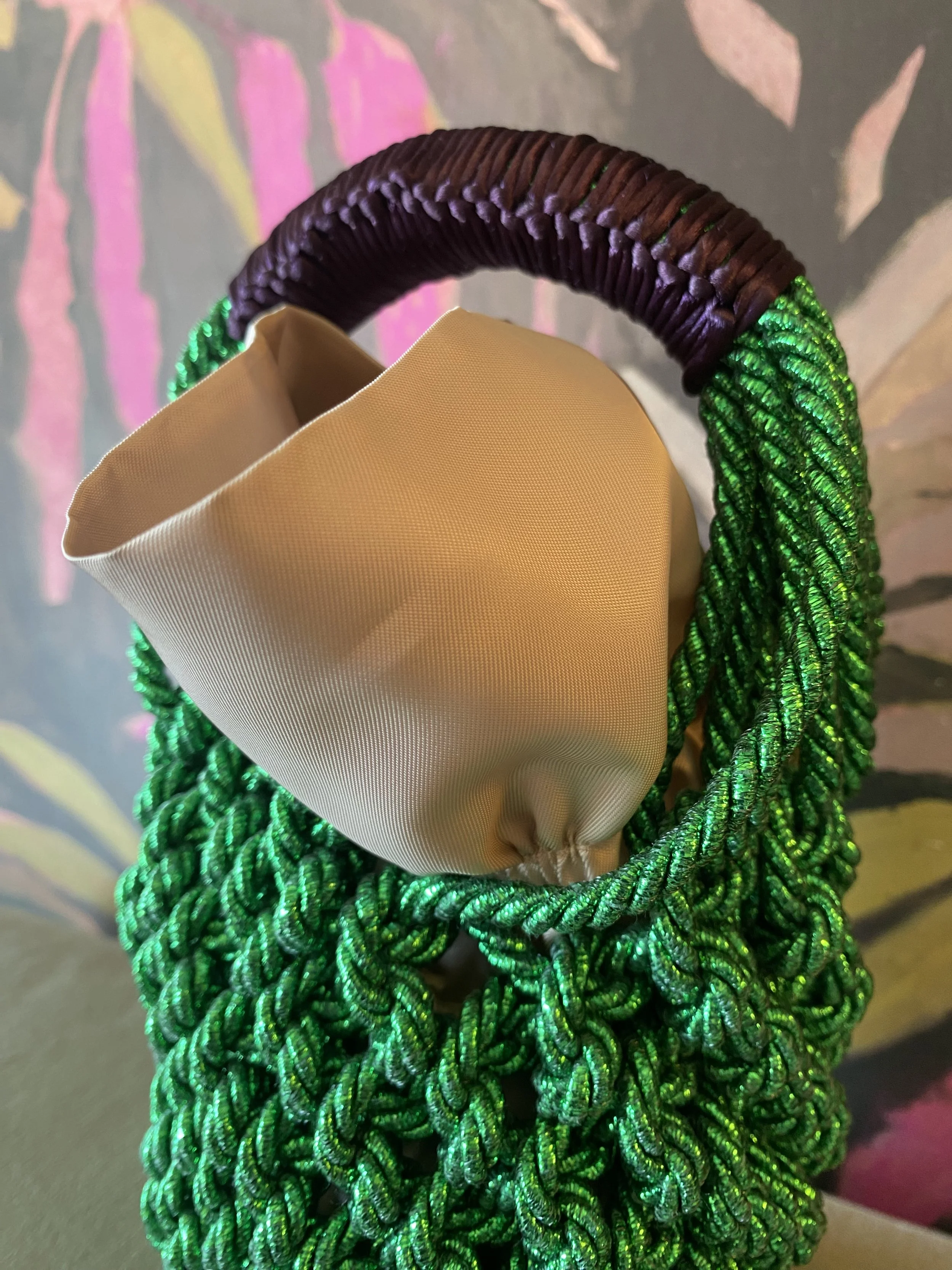 Borsa con manico avvolto in filato verde e manico in fibra marrone, con fusione beige all’interno, sfondo con motivo floreale rosa e verde.