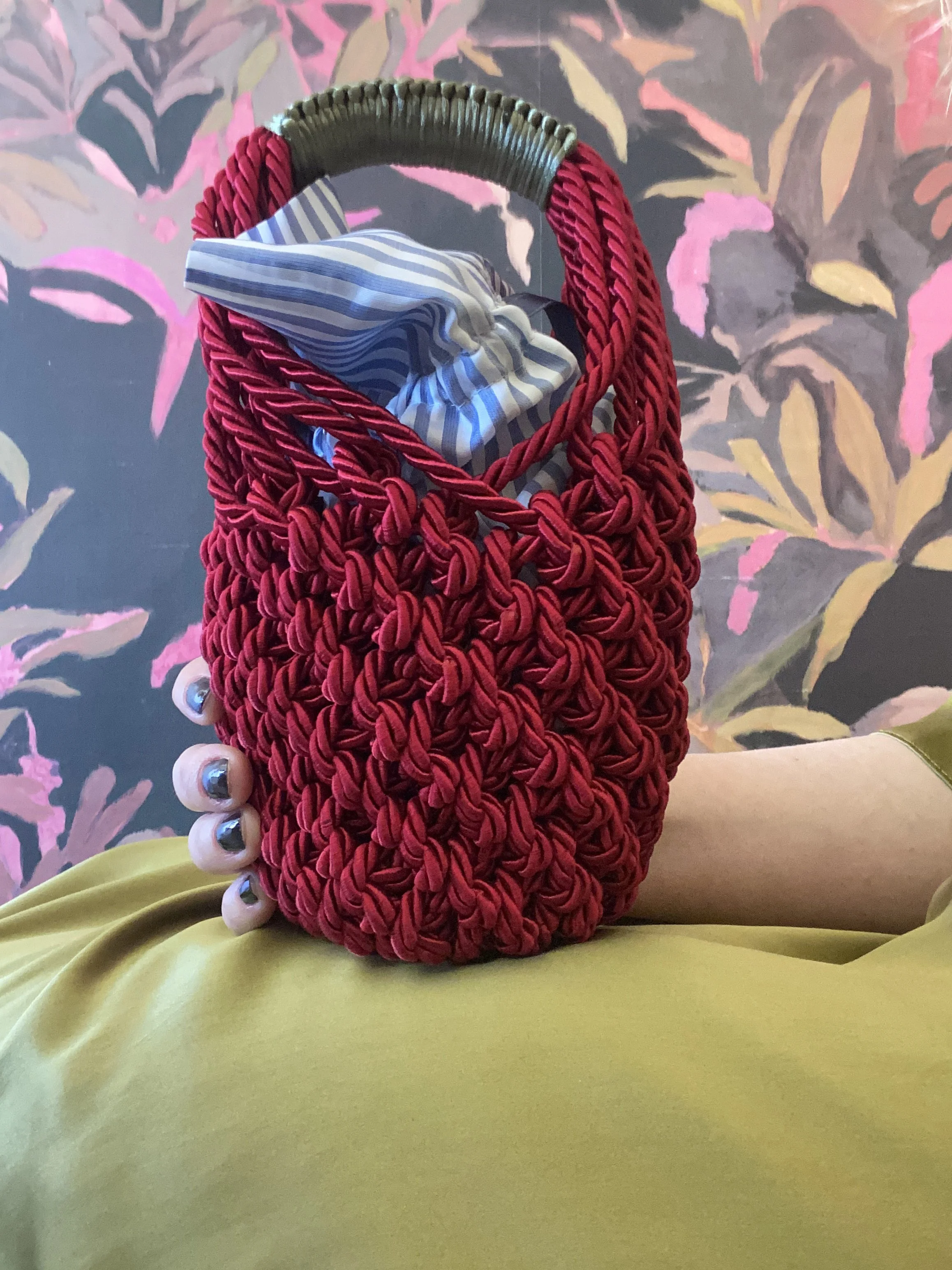Borsa rossa intrecciata con manico in legno, contenente una tasca a righe bianche e blu, su sfondo con motivo floreale rosa e grigio.
