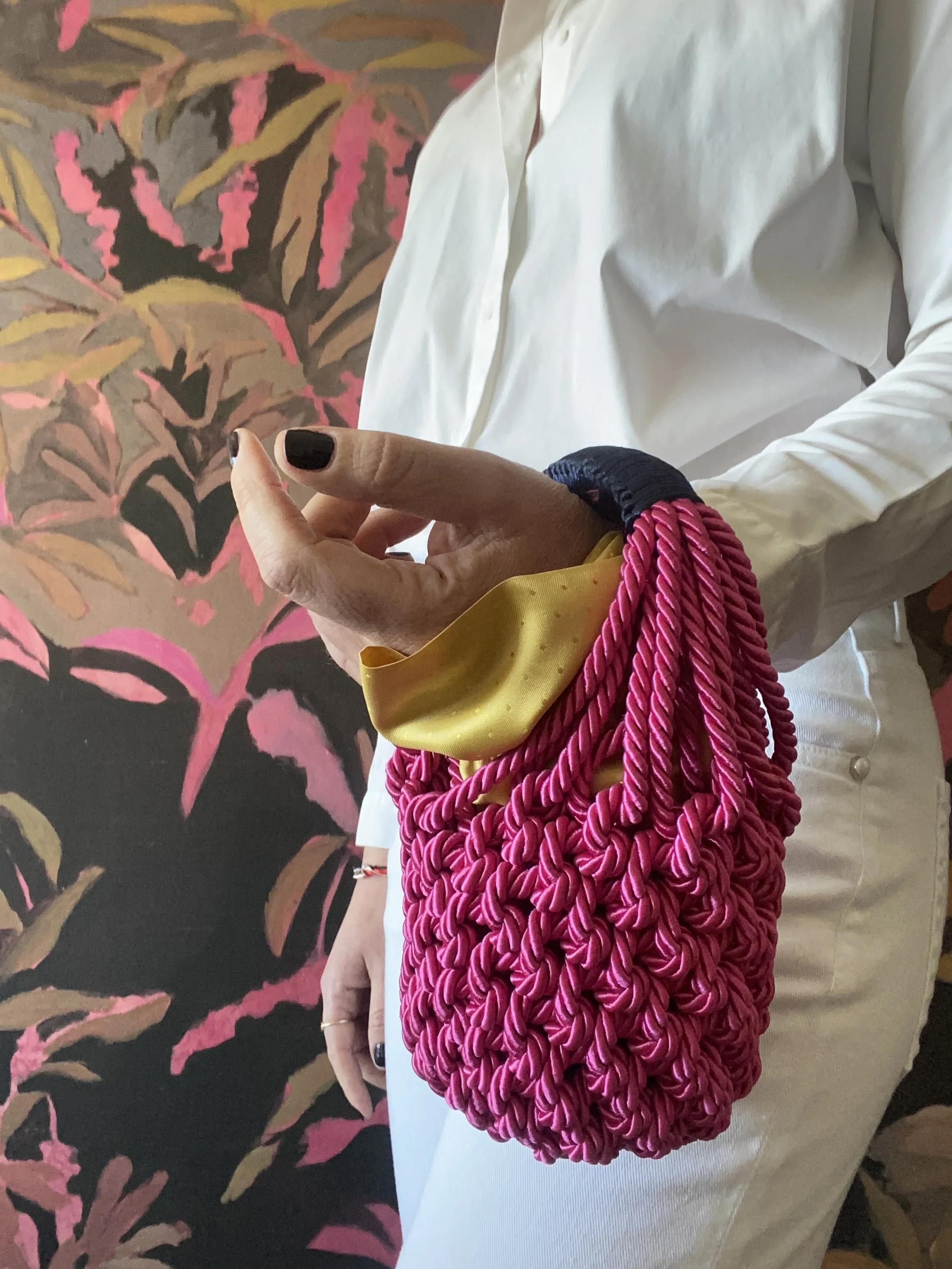 Una mano tiene una borsa color rosa con una chiusura gialla e il polso avvolto da una manica di velluto blu e rosa, con sfondo di tessuto con pattern di foglie rosa e marroni.