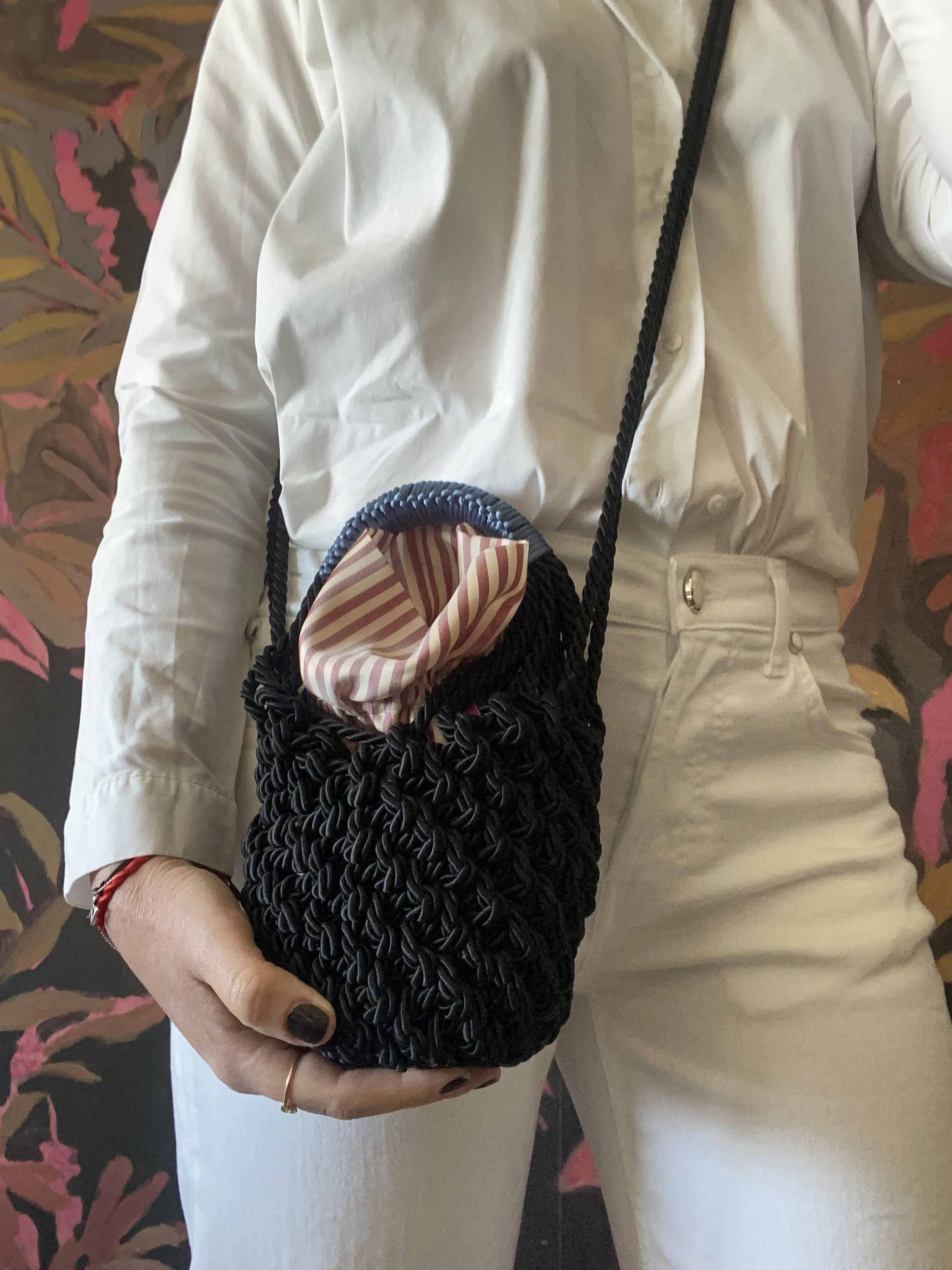 Una persona con abbigliamento bianco e pantaloni bianchi mostra una borsetta nera fatta intrecciata a mano con una fodera a righe rosse e bianche, con una tracolla nera.