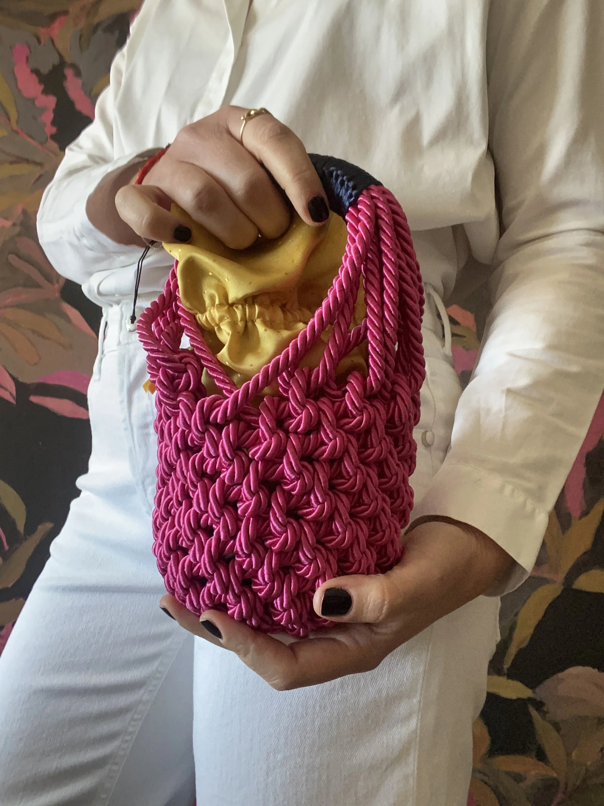 Una persona che indossa un abito bianco con le mani dipinte di nero, tiene in mano una borsa rosa fatta a maglia con una fodera gialla.