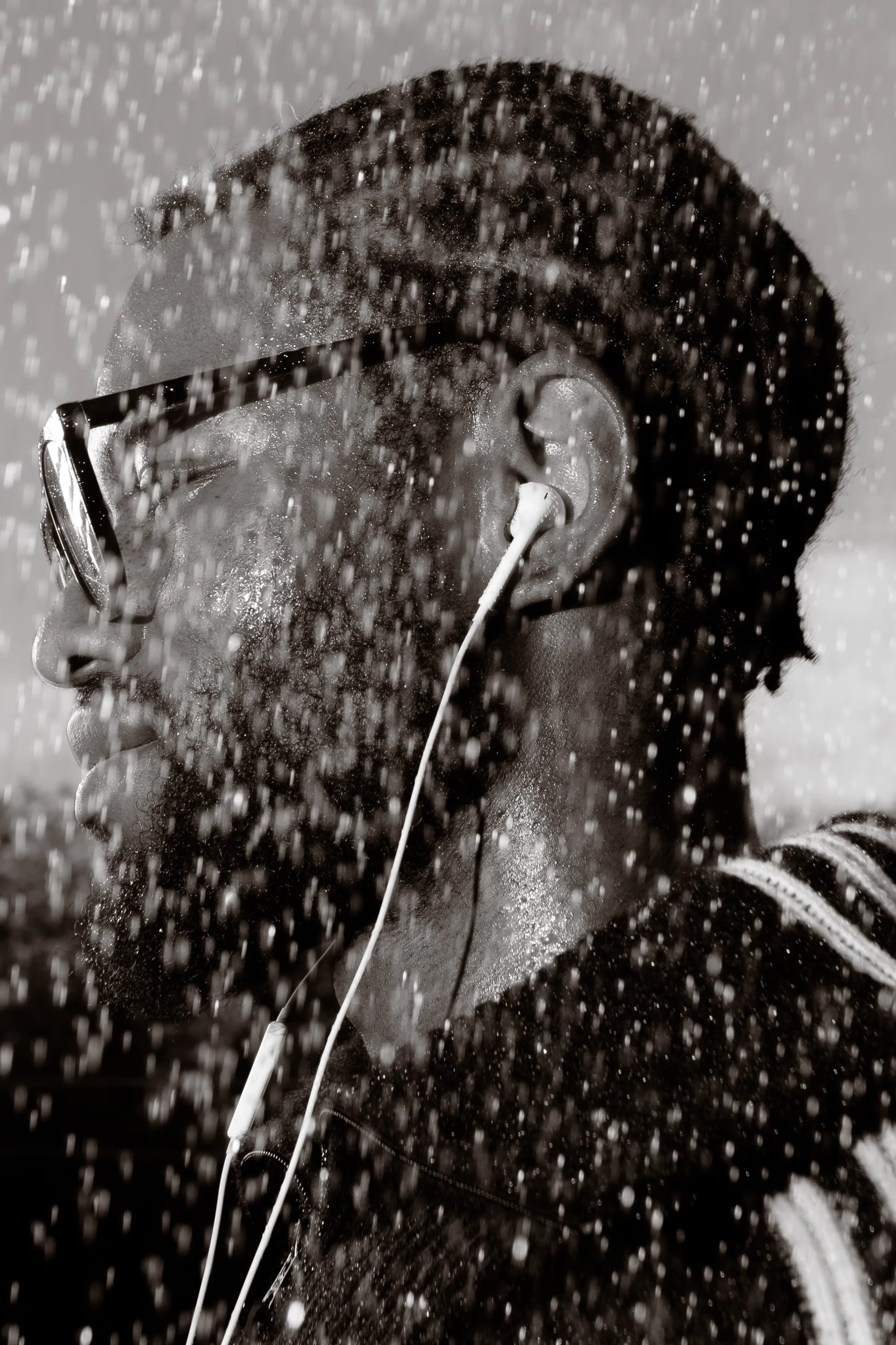 Black and white close-up portrait of a man wearing sunglasses and wired earphones in heavy rain, photographed by Yaroslav Prytula for Common Frequency at Factory International in Manchester.
