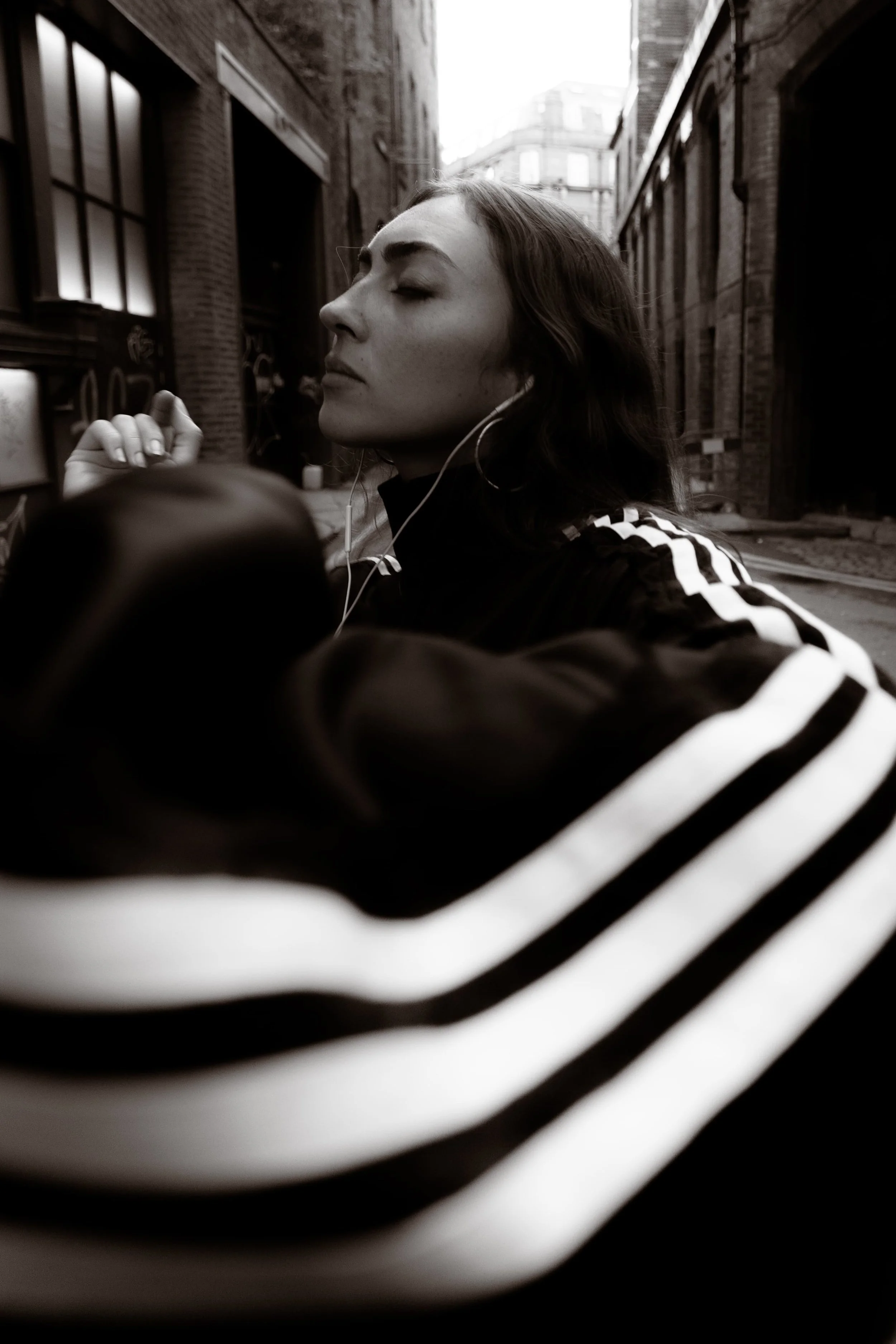 Black and white portrait of a woman wearing an adidas tracksuit and wired earphones in an industrial Manchester setting, photographed by Manchester and London based photographer Yaroslav Prytula for Common Frequency.