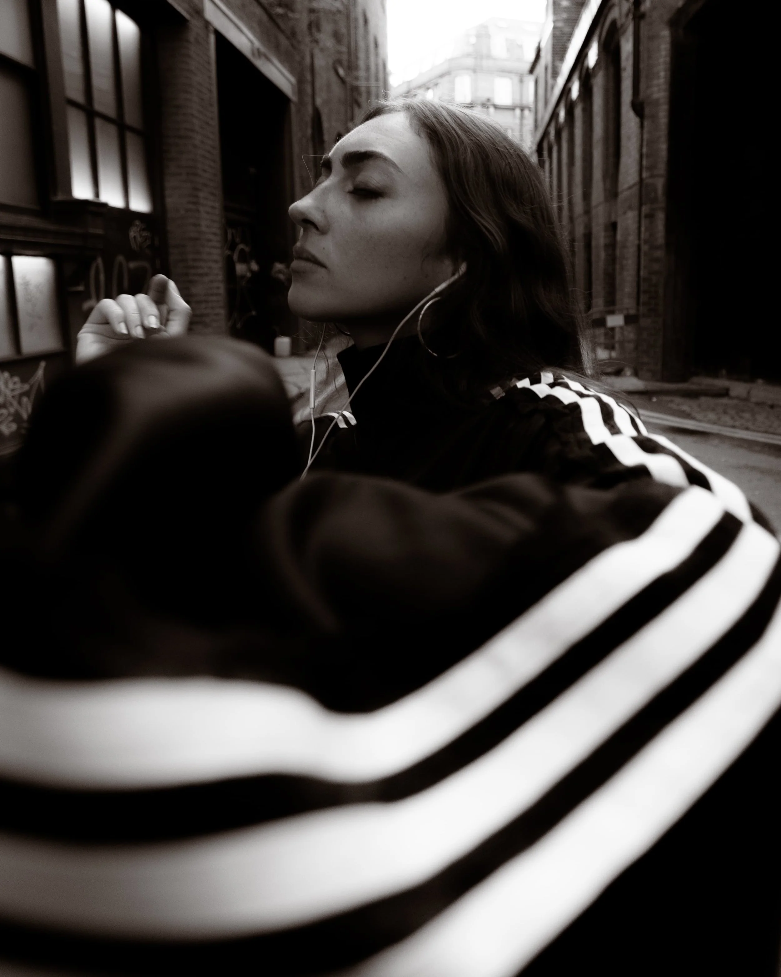 Black and white portrait of a woman wearing an adidas tracksuit and wired earphones in an industrial Manchester setting, photographed by Manchester and London based photographer Yaroslav Prytula for Common Frequency.