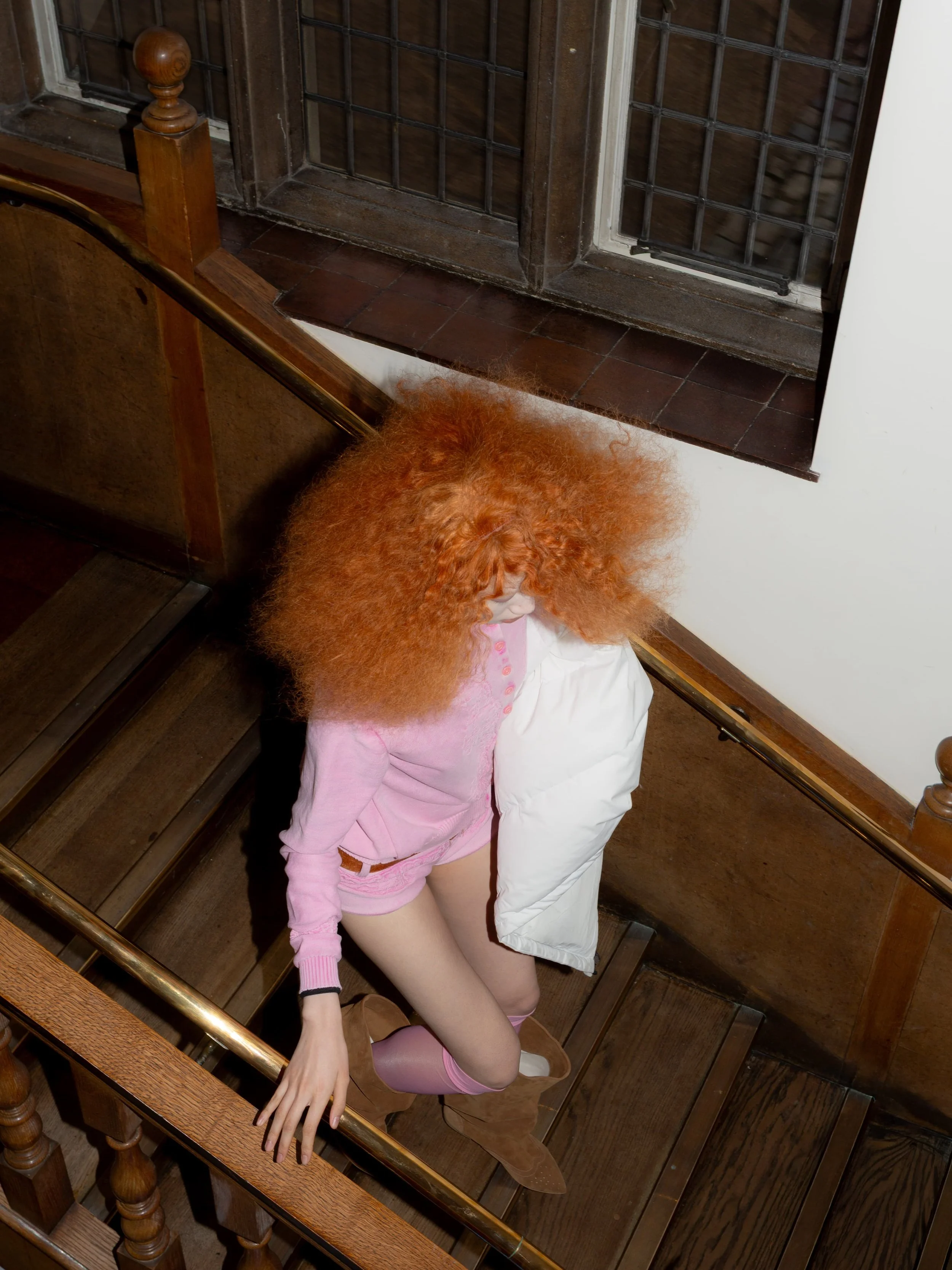 Model with voluminous red hair wearing a pink knitted outfit and white padded garment on a staircase backstage at Masha Popova AW26.