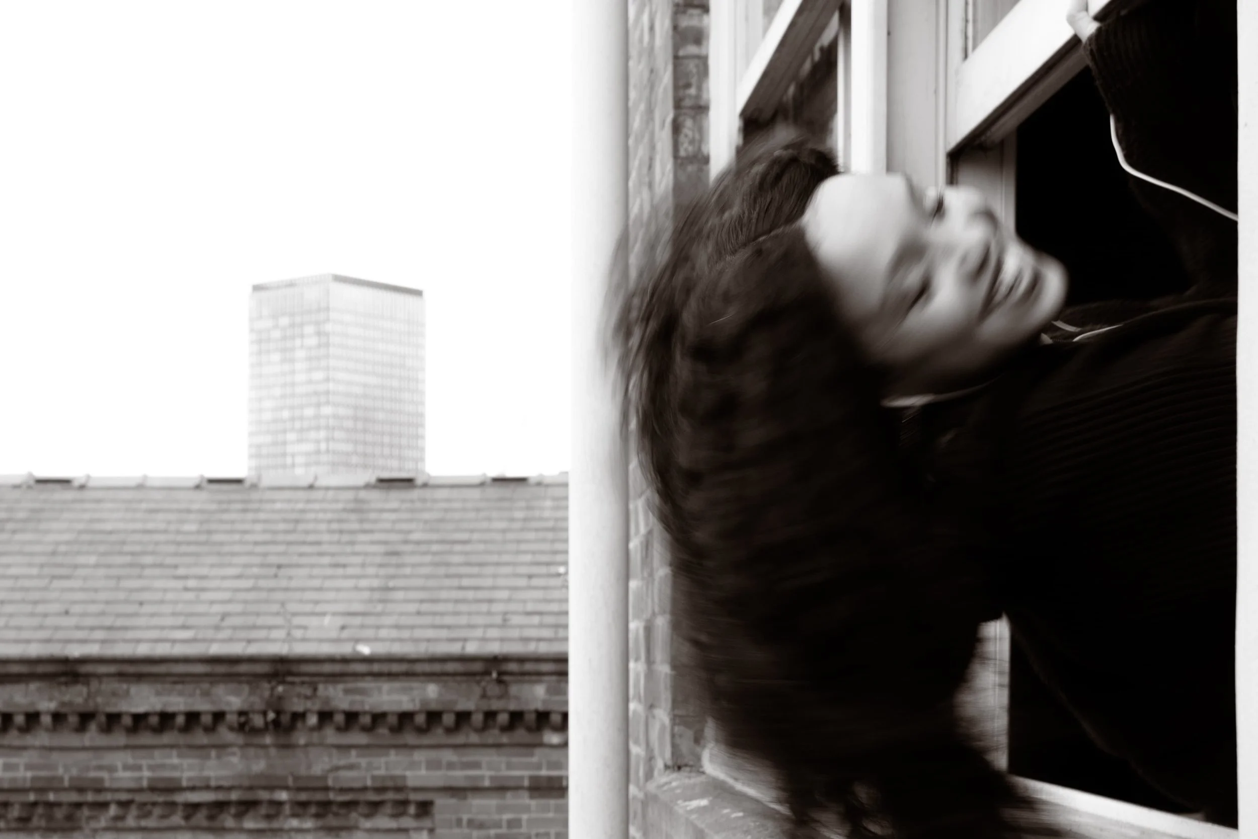 Black and white photograph of a blurred figure leaning through a window overlooking an industrial Manchester cityscape, photographed by Yaroslav Prytula for Common Frequency at Factory International.