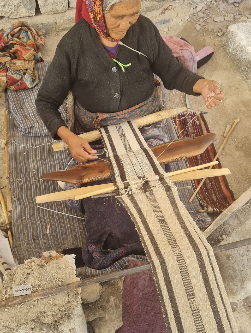 An elderly woman wearing a red headscarf and black sweater is sitting on a striped mat, weaving a fabric with traditional patterns using a backstrap loom in an outdoor setting.