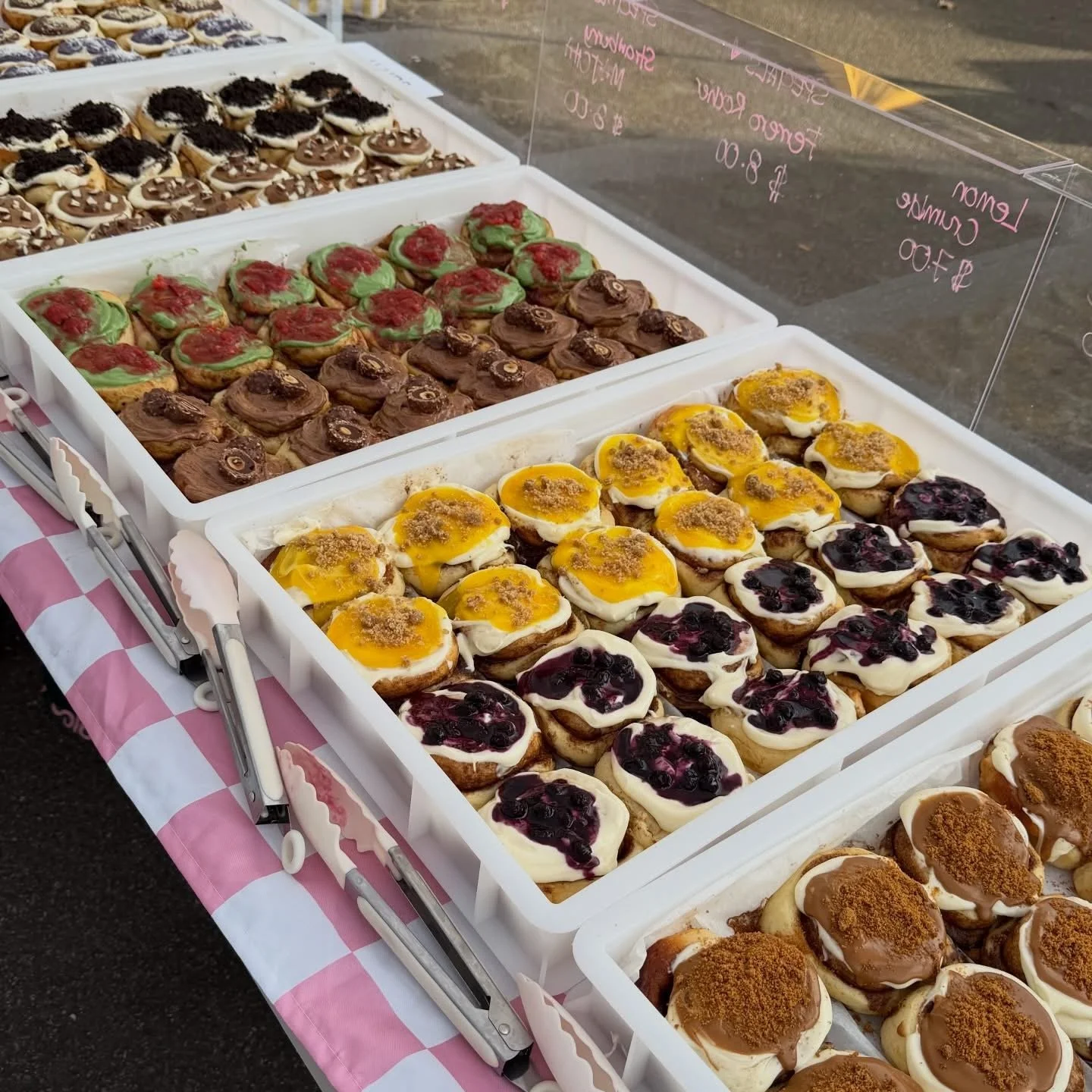 A display of assorted donuts at an outdoor market, including chocolate, green icing with red topping, lemon glaze with crumb topping, and blueberry toppings.