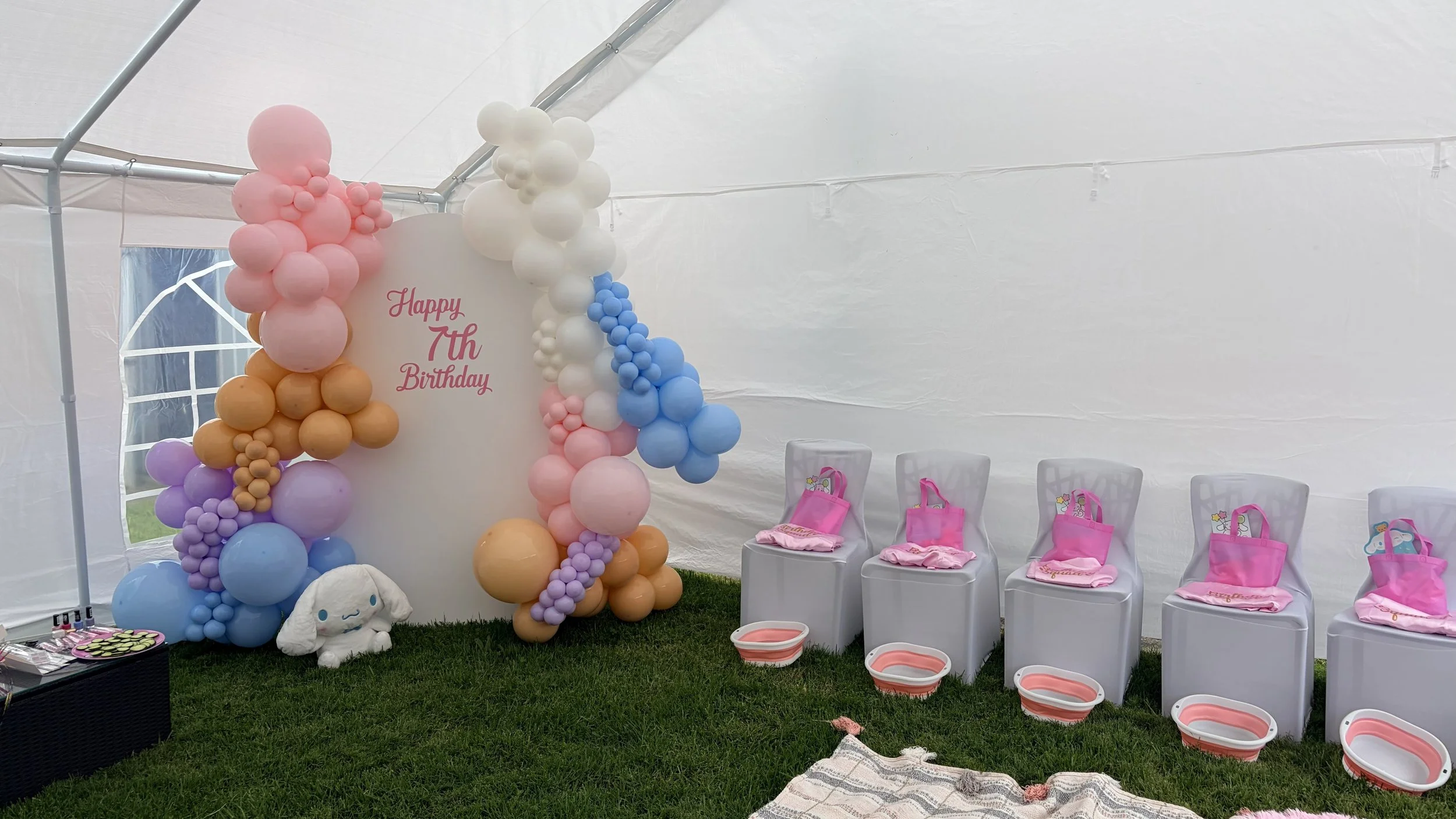 Decorated area for a child's birthday party with pink, purple, white, peach, and blue balloons forming an arch, chairs with pink party bags and towels, and a small plush bunny with a unicorn horn, inside a white tent with grass underneath.