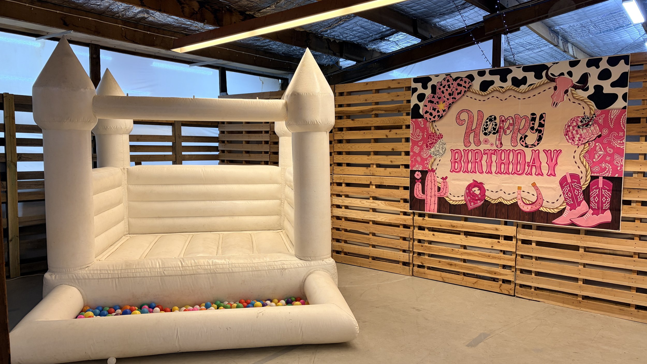 White inflatable castle with tall turrets inside a wooden-fenced area, next to a colorful birthday sign that says "Happy Birthday" with cowboy-themed decorations.