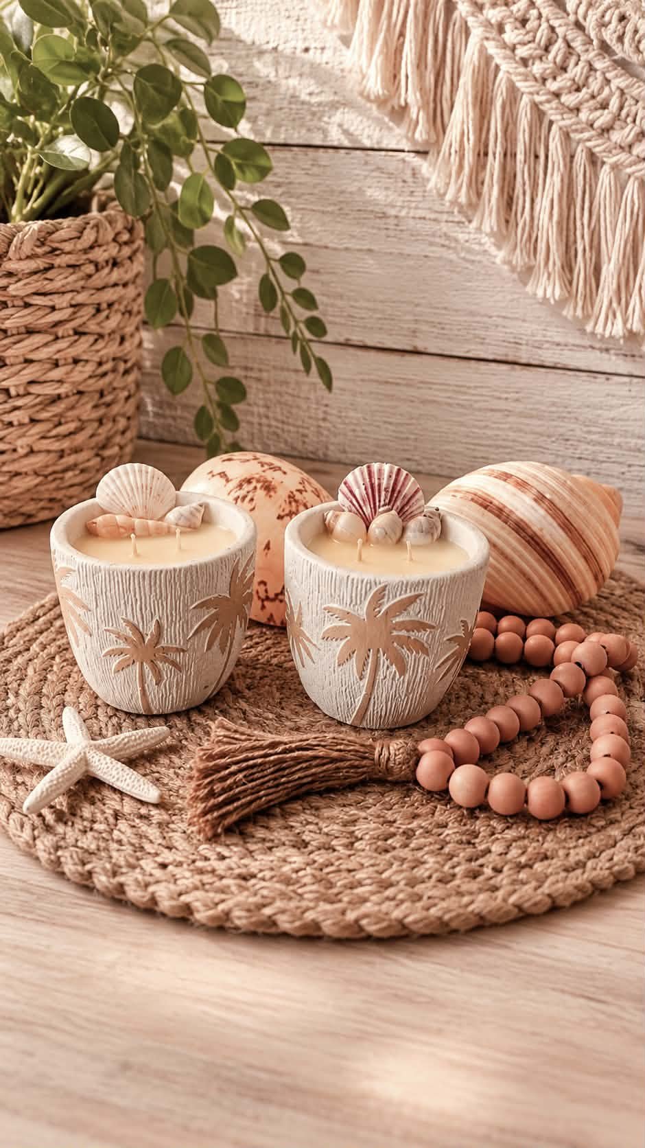 Decorative seaside scene with two white candles featuring palm tree carvings, surrounded by seashells, a starfish, a brown tassel, a pink beaded necklace, a large shell, and a green plant in a woven basket, set on a woven circular mat against a white wood-paneled background with a beige woven wall hanging.
