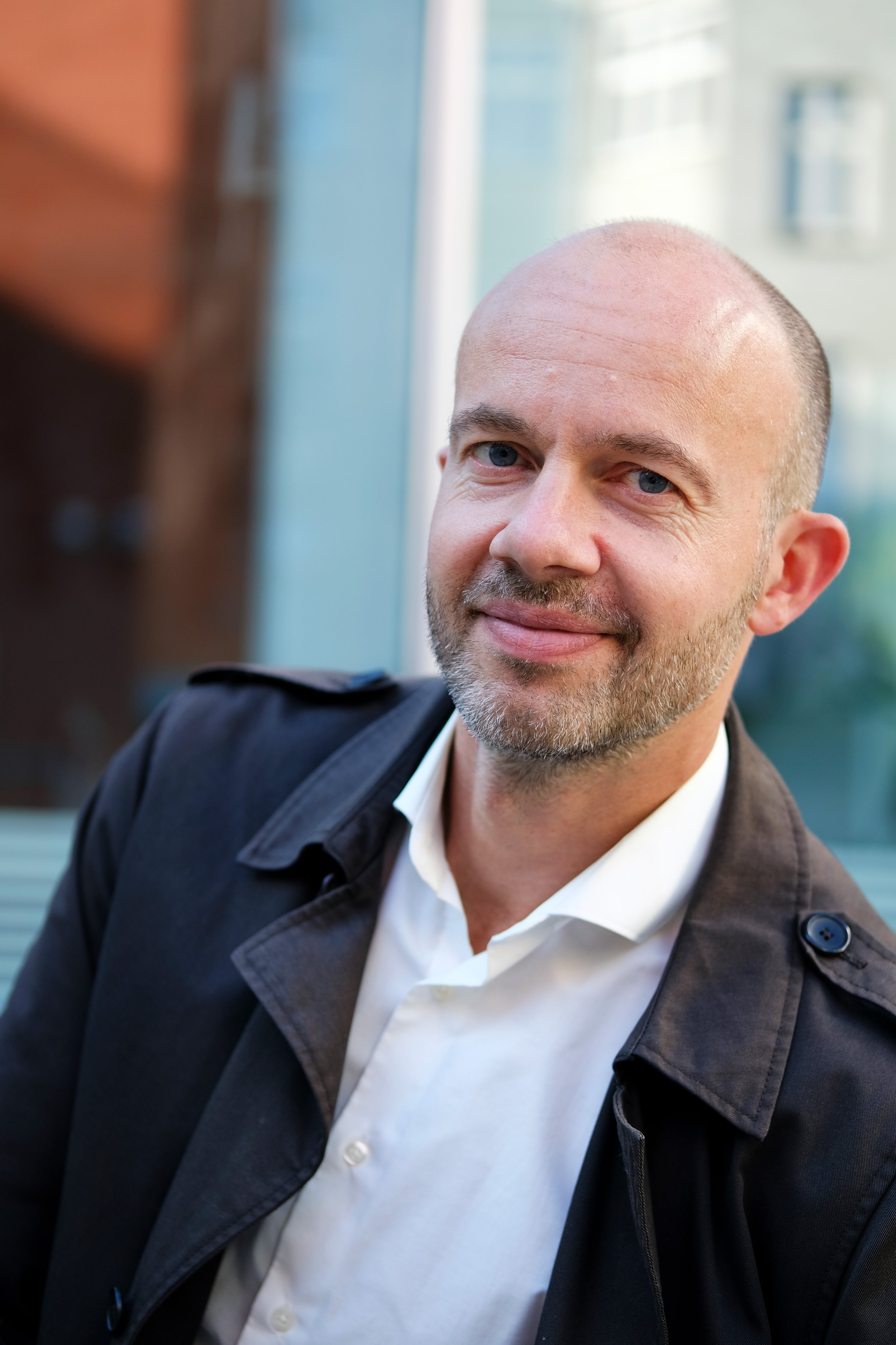 Bald man with a beard wearing a black coat and white shirt, smiling outdoors.