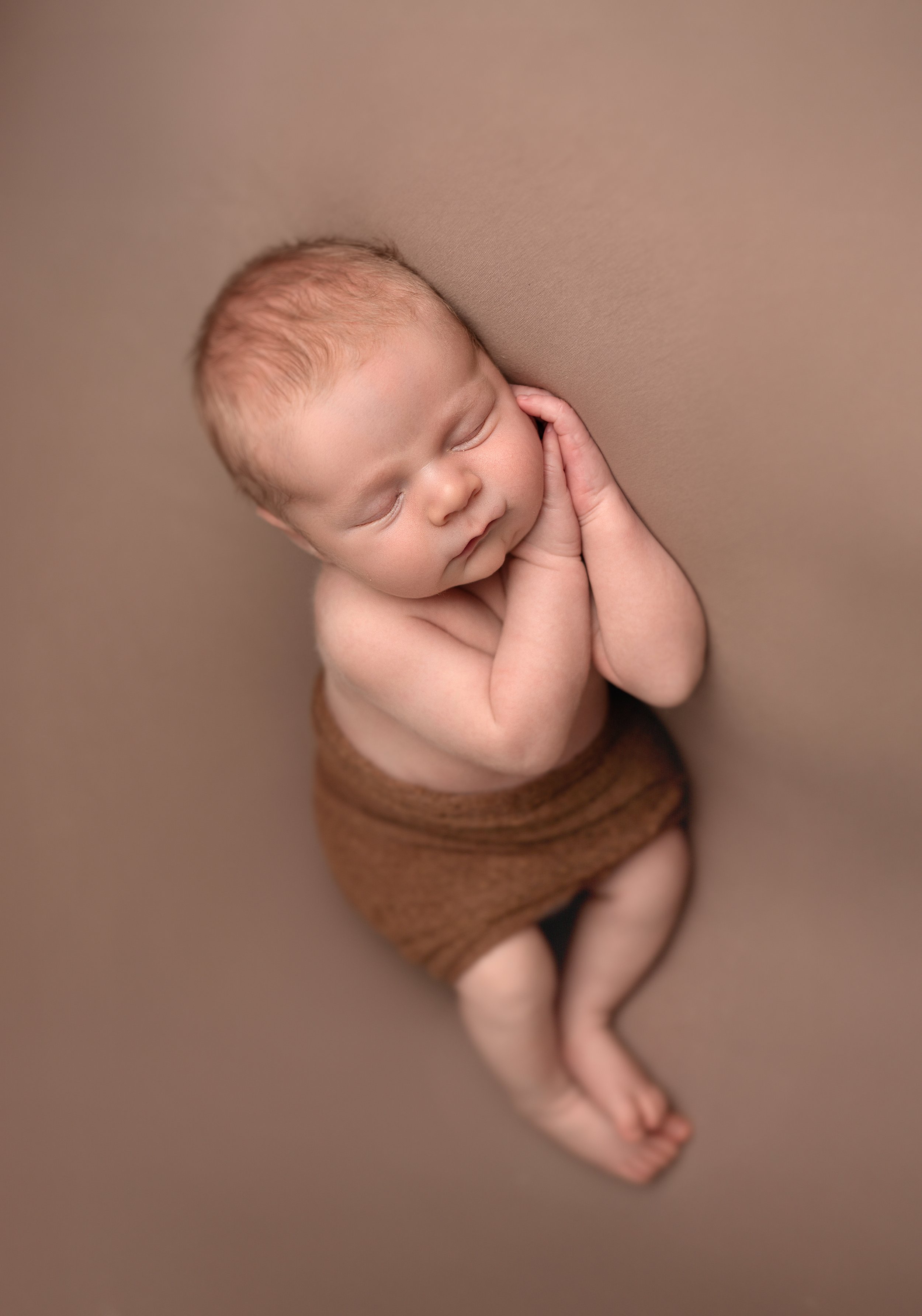 A sleeping baby with curly hair, resting with hands near face, wearing a brown wrap around the waist, lying on a soft beige surface. Best newborn, family and kids photographer in Northamptonshire, Bedfordshire, Buckingamnshire. Timeless