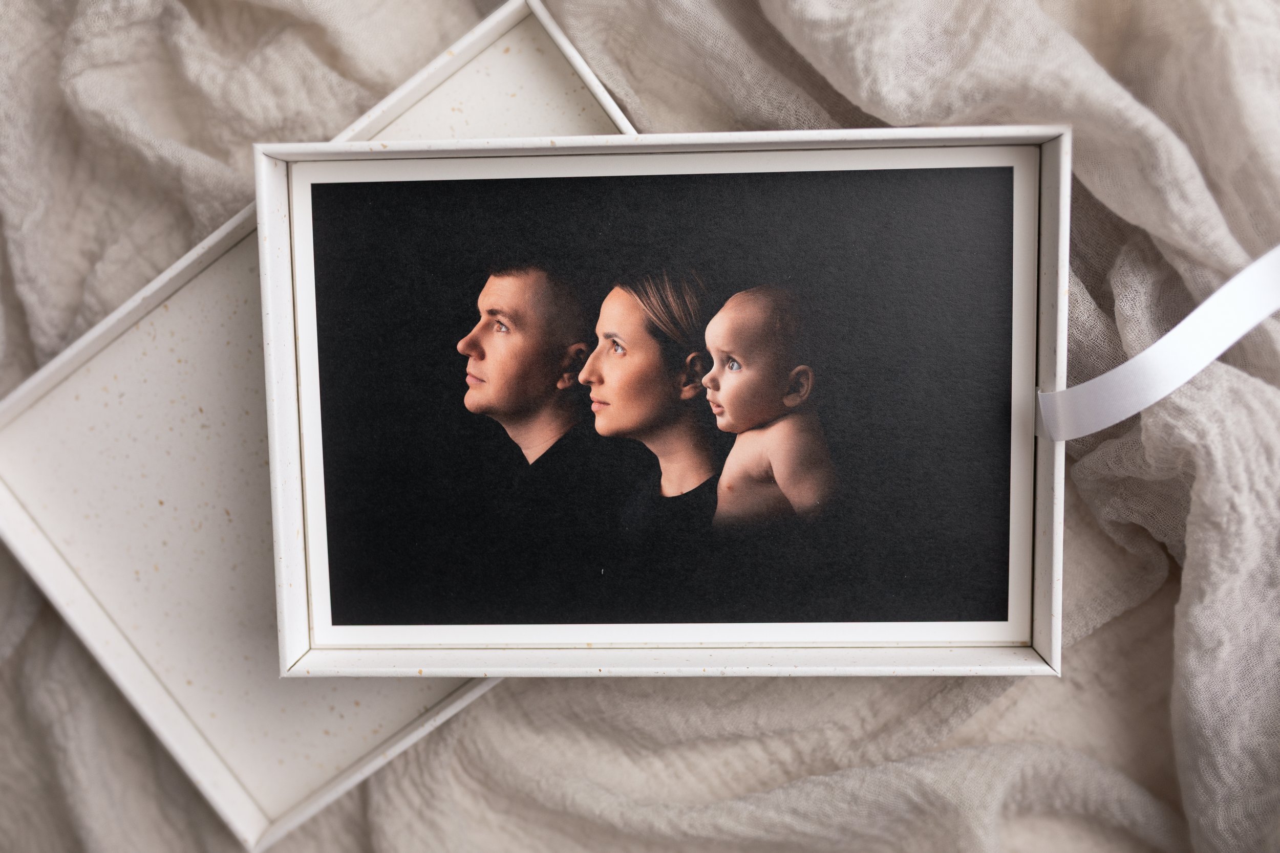 A framed photograph of a family showing a man, woman, and baby in profile against a black background, placed on beige fabric.