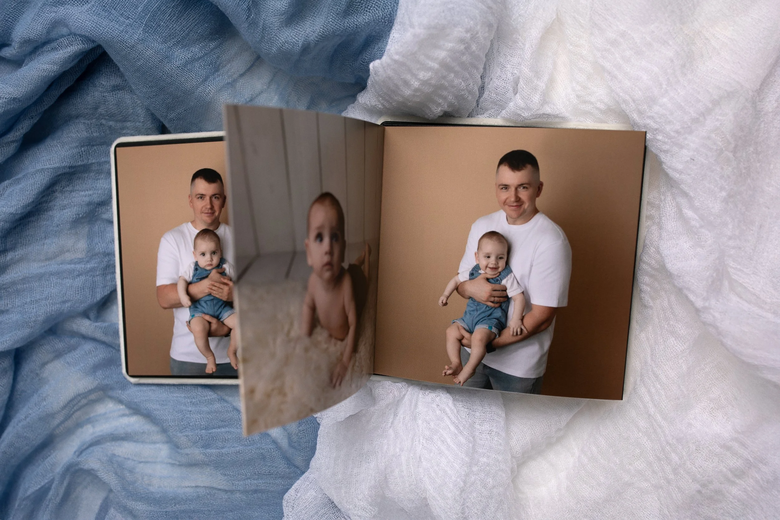 Color photograph of a family  including a father, and one baby boy, posed together and smiling. The photo is set against a neutral background.