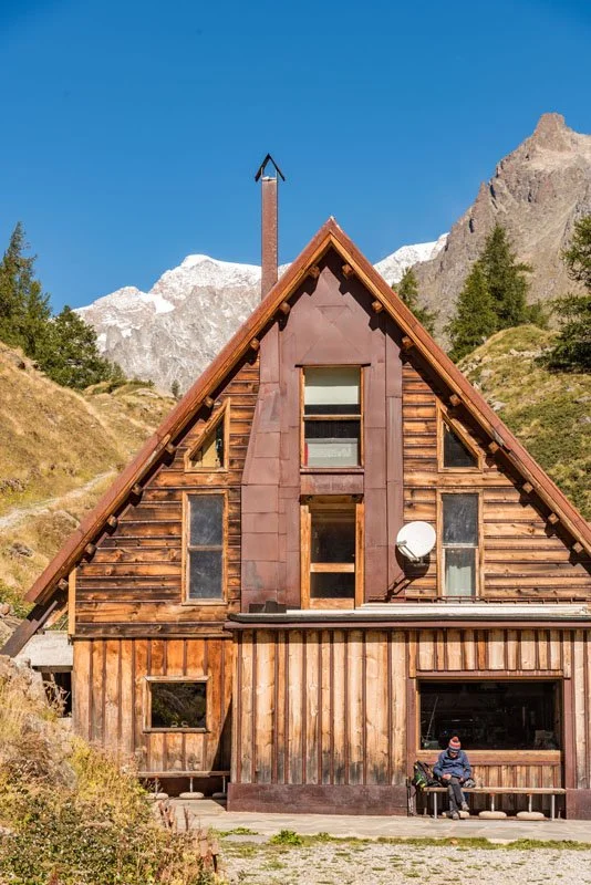 A rustic A-frame wooden house in a mountainous setting with trees and snow-capped peaks in the background, with a person sitting on a bench outside.