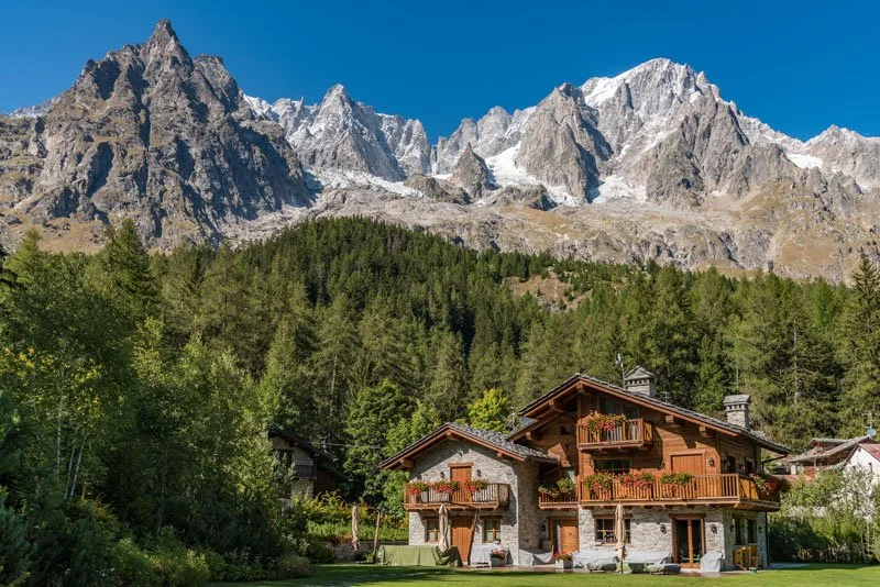 A mountain landscape with rocky peaks in the background, dense green forest in the middle, and traditional mountain houses in the foreground.