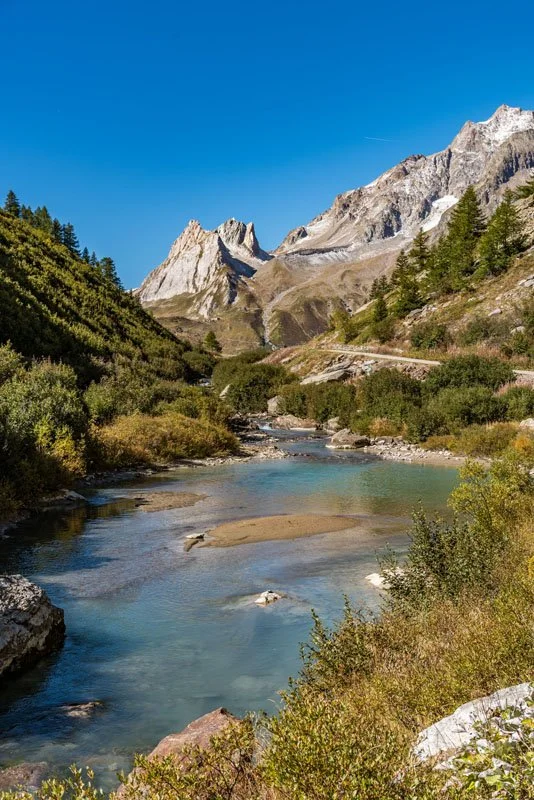 Scenic mountain landscape with a river flowing through green shrubbery and rocky terrain under a clear blue sky.