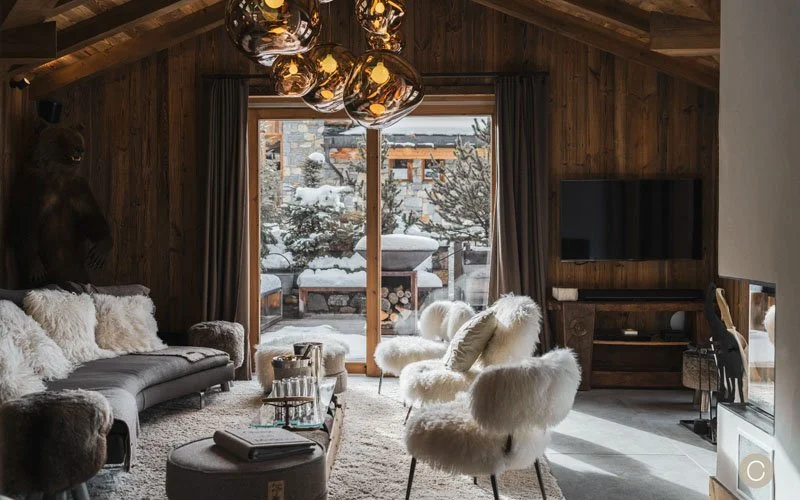 A cozy living room with wooden walls and high ceiling, featuring a gray sofa with fur pillows, two fur-covered chairs, a glass coffee table, a large flat-screen TV, and a sliding glass door opening to a snowy outdoor scene with trees and stacked logs.