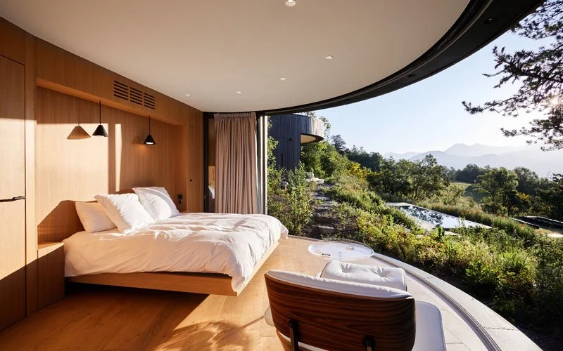 A modern hotel room with a large bed, wooden paneling, and a curved floor-to-ceiling window with a view of greenery and mountains.