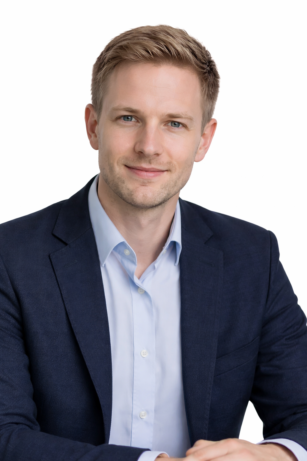 Portrait of a young man with blonde hair and blue eyes wearing a navy blazer and light blue dress shirt, smiling softly, against a white background.
