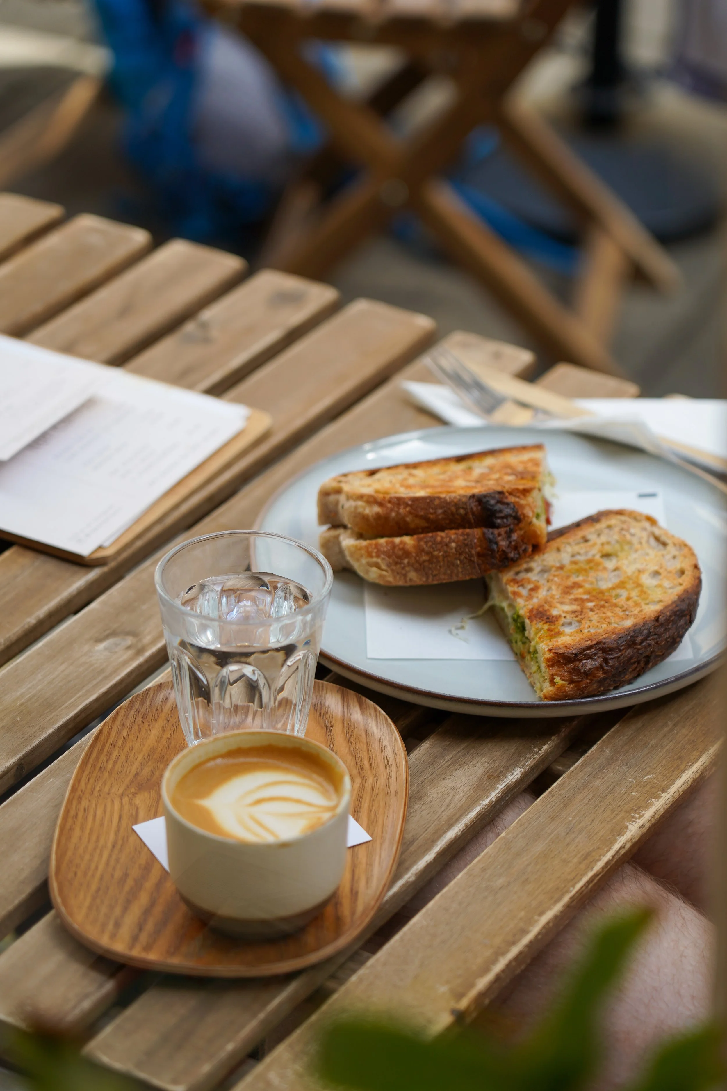 A wooden table set with a glass of water, a cappuccino with latte art on a wooden tray, and a plate with two toasted sandwiches.