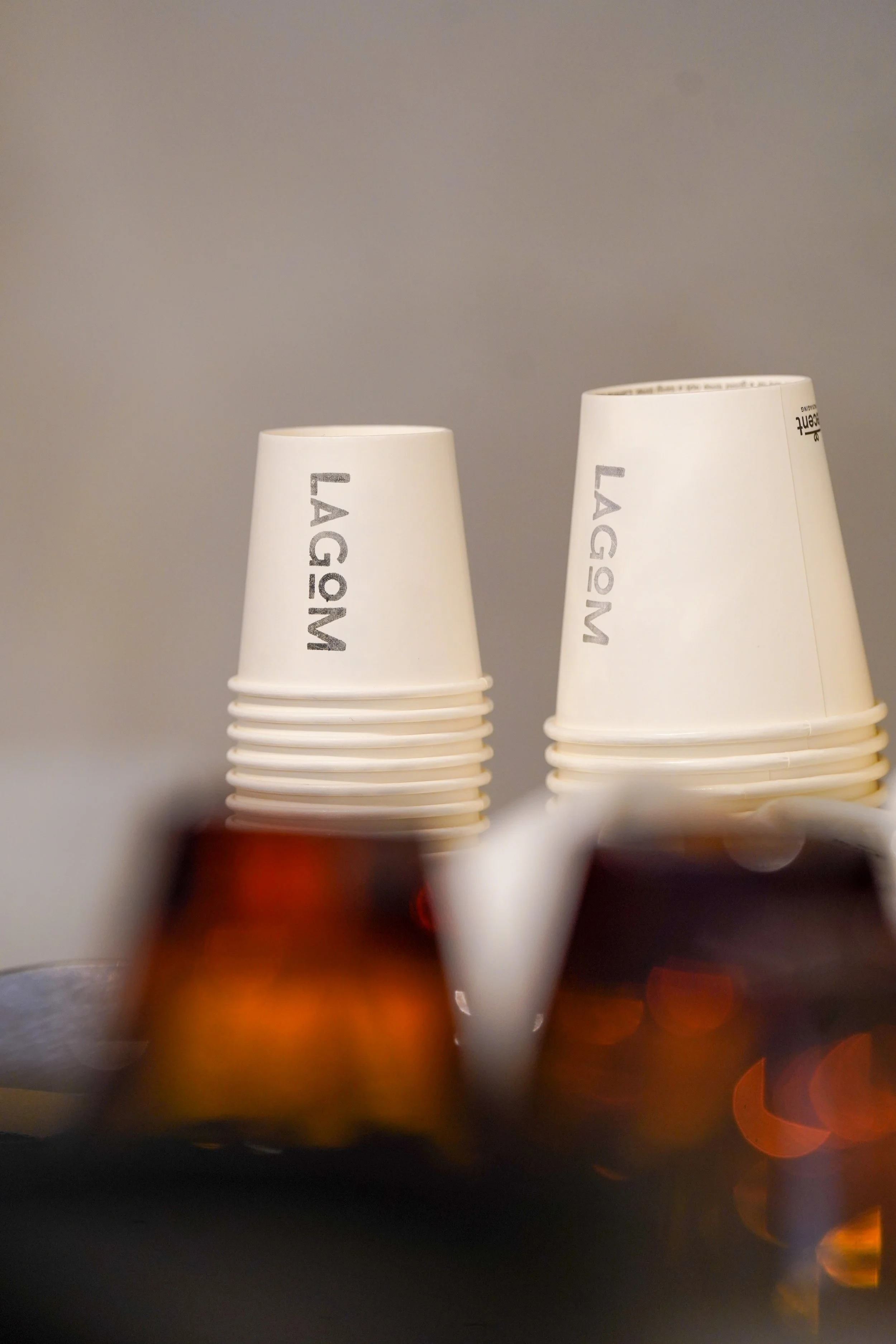 Two stacks of white paper cups with "LAGOM" printed on them, placed on a dark surface with blurred objects in the foreground.