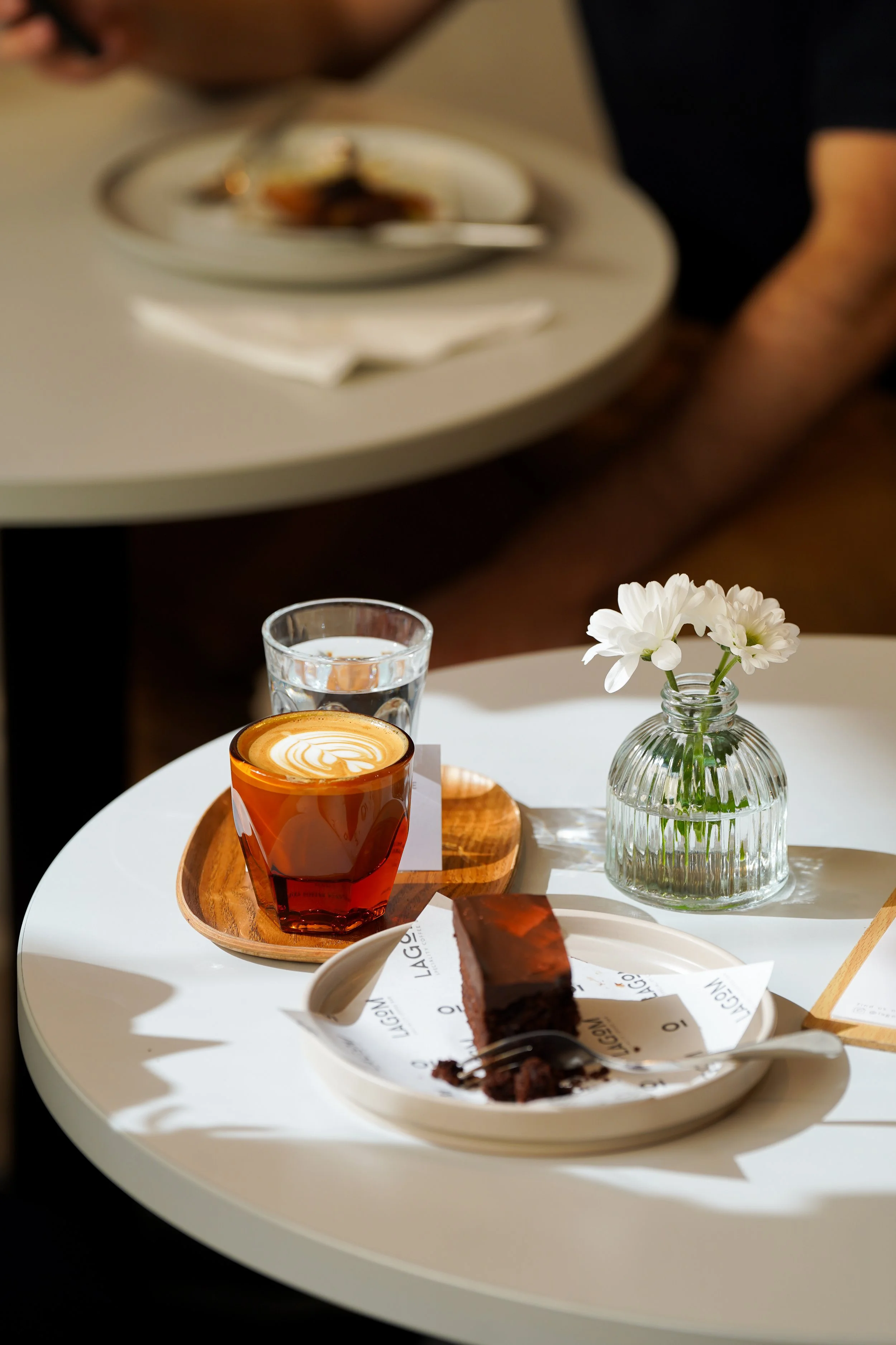 A white table with a glass of water, a cup of coffee with latte art, a small vase of white flowers, and a plate with a slice of chocolate cake.