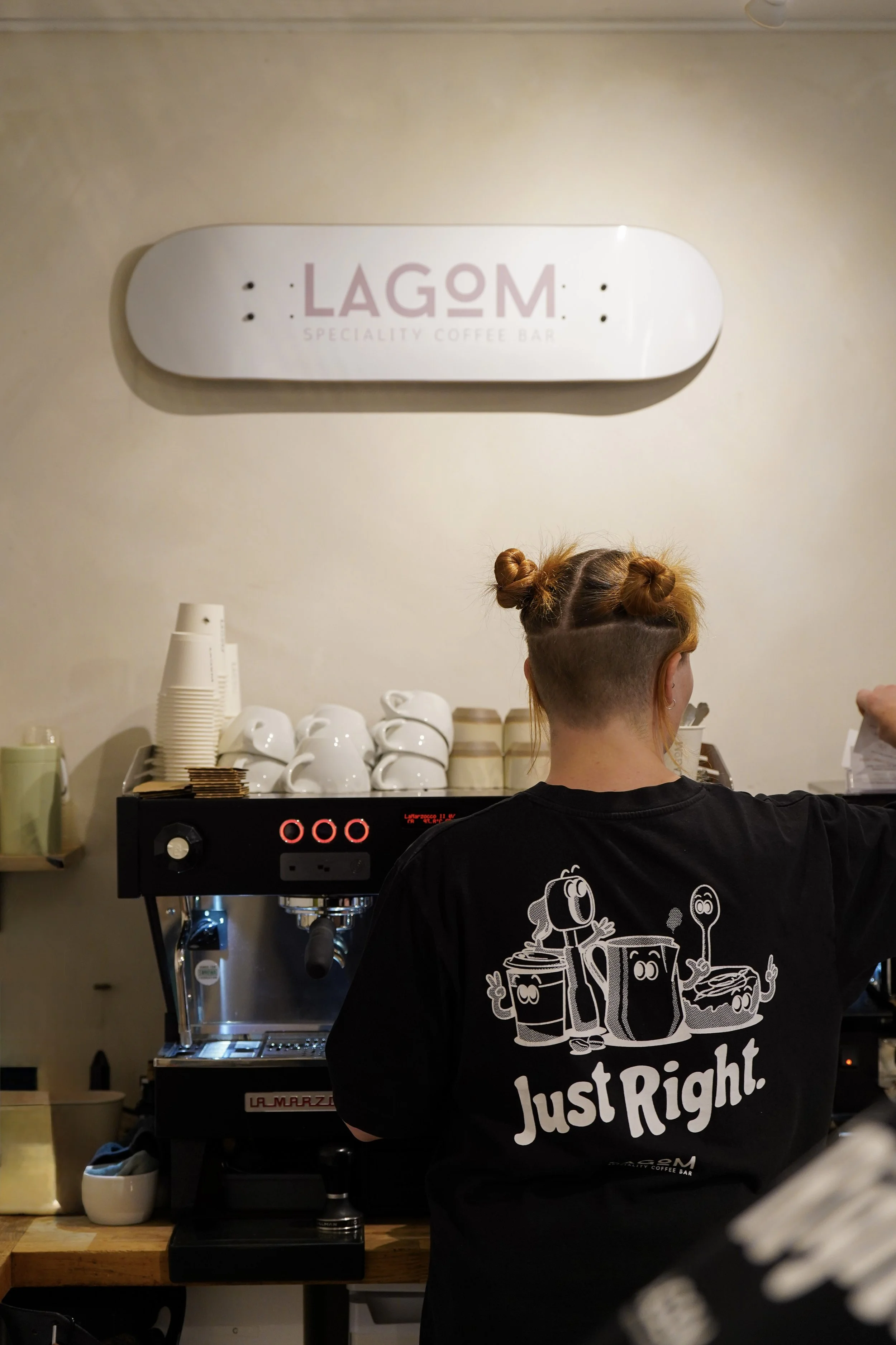 A barista at a coffee shop behind an espresso machine, with cups and small containers on the counter. A sign on the wall reads 'LAGON Specialty Coffee Bar'. The person is wearing a black T-shirt with a graphic of a coffee cup character and the words 'Just Right.'